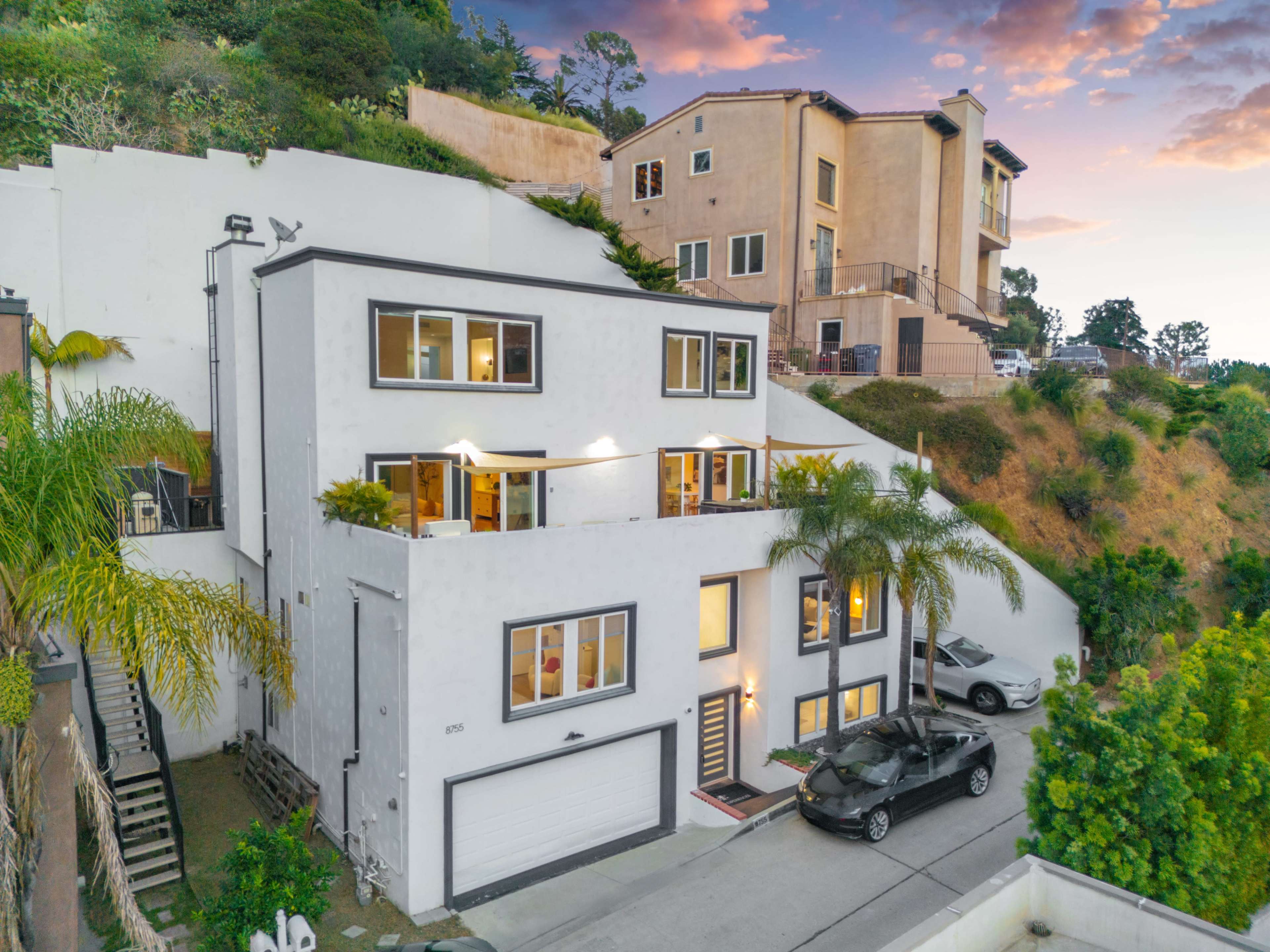 The image shows a modern two-story house situated on a hillside, with a driveway and several palm trees in the foreground.