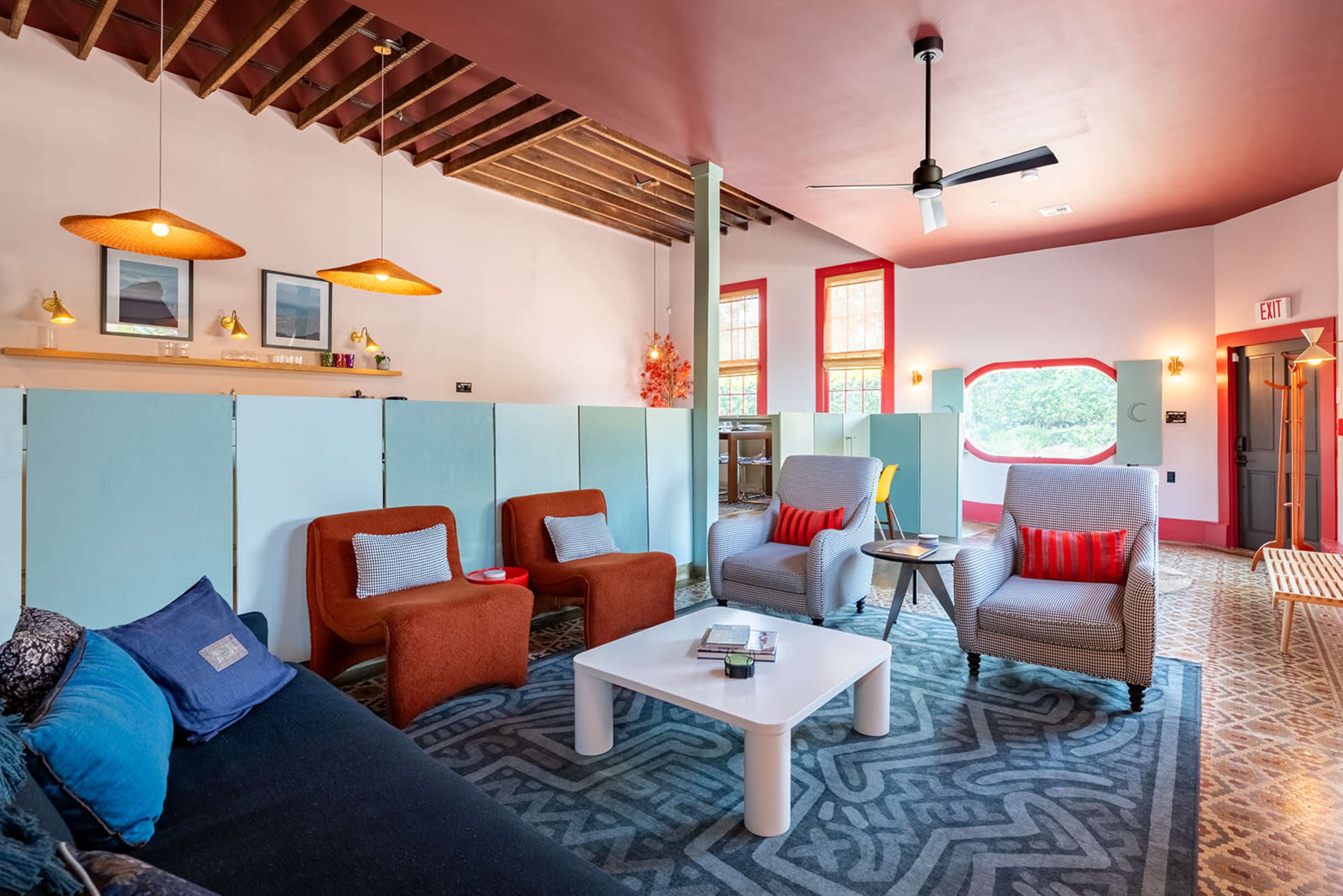 The image shows a brightly colored lounge area with a mix of seating options, including chairs and a sofa, under decorative lighting and wooden beams.