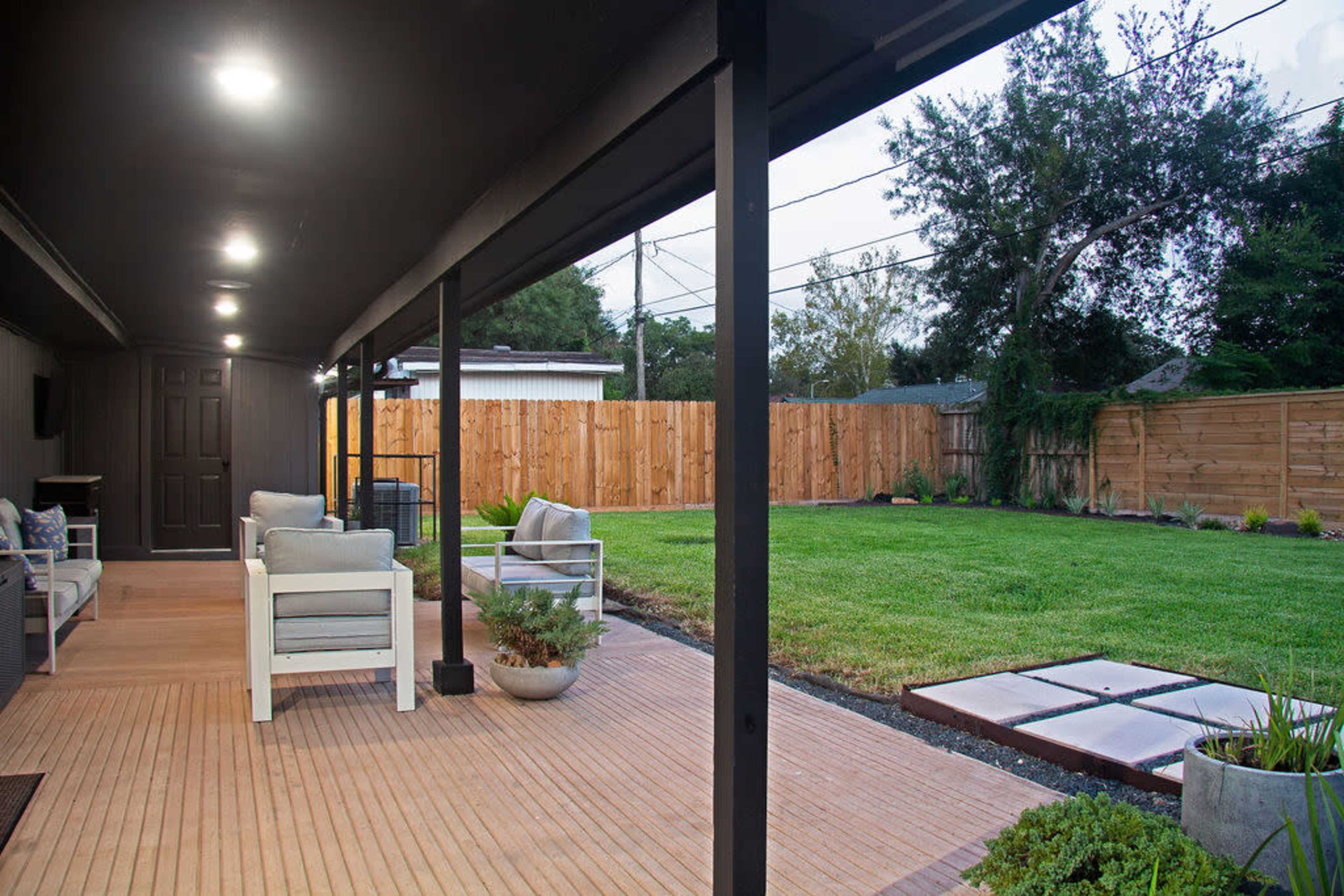 A covered patio with seating arrangements overlooking a grassy yard enclosed by a wooden fence.