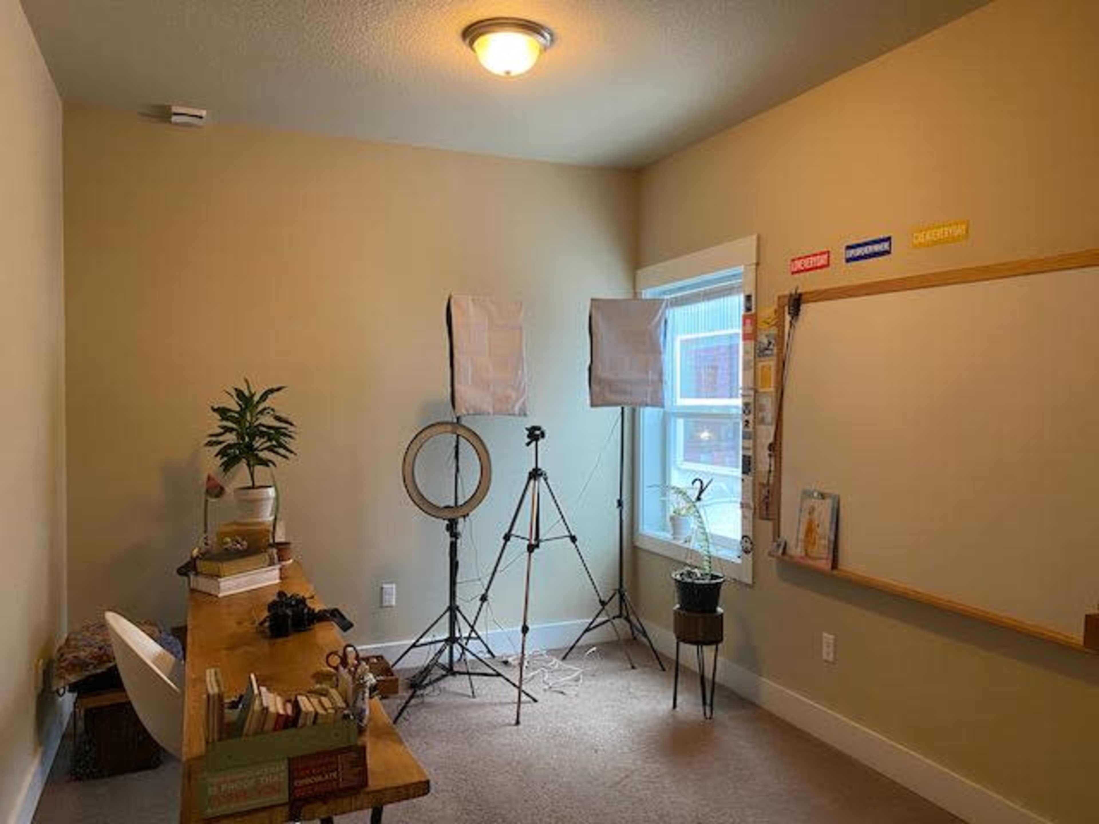 A well-lit room features a desk with supplies, a circular mirror, two light stands, and a whiteboard against the wall.