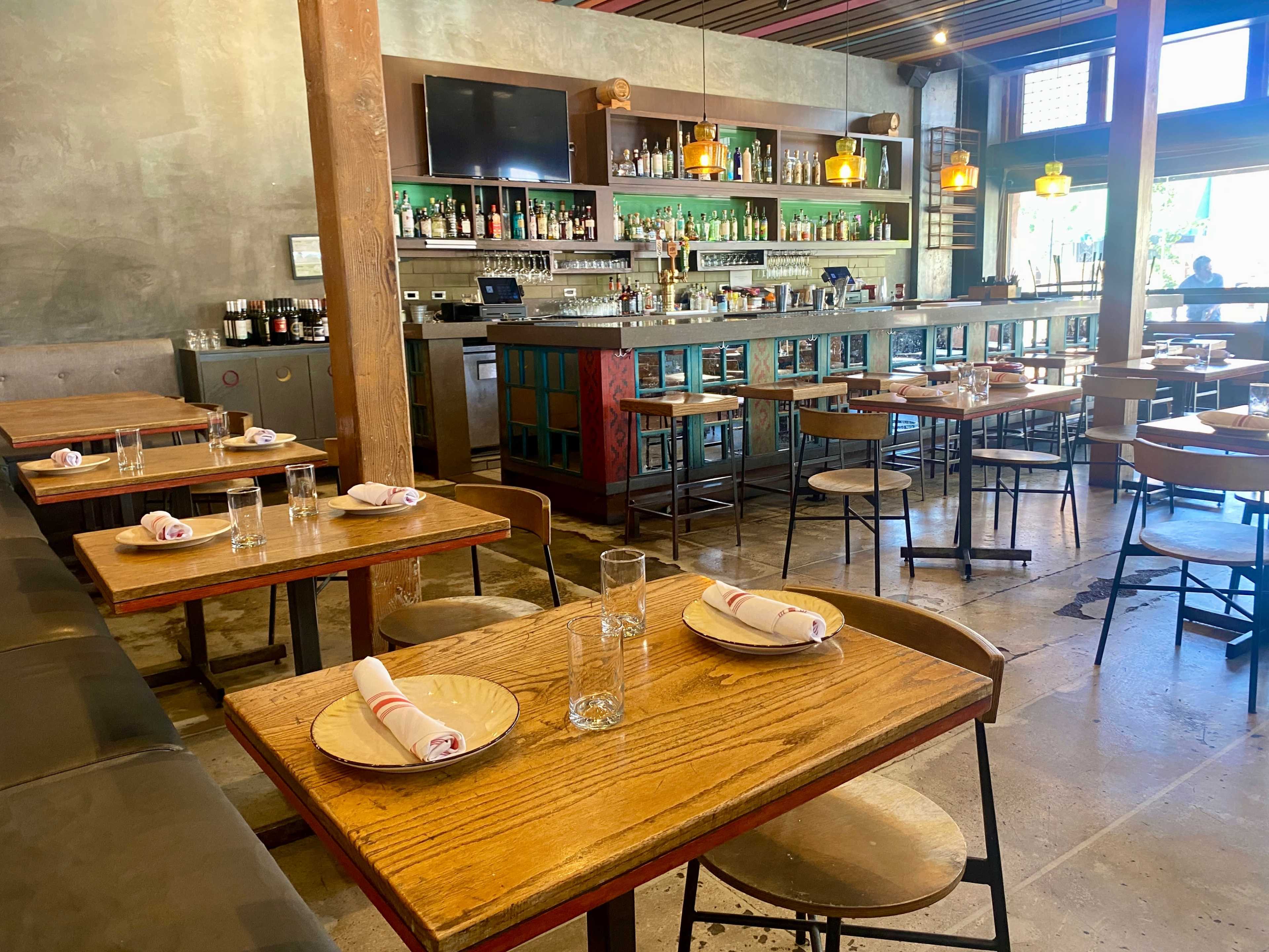 A restaurant interior featuring wooden tables set with plates and napkins, a bar area stocked with liquor, and large windows allowing natural light to enter.
