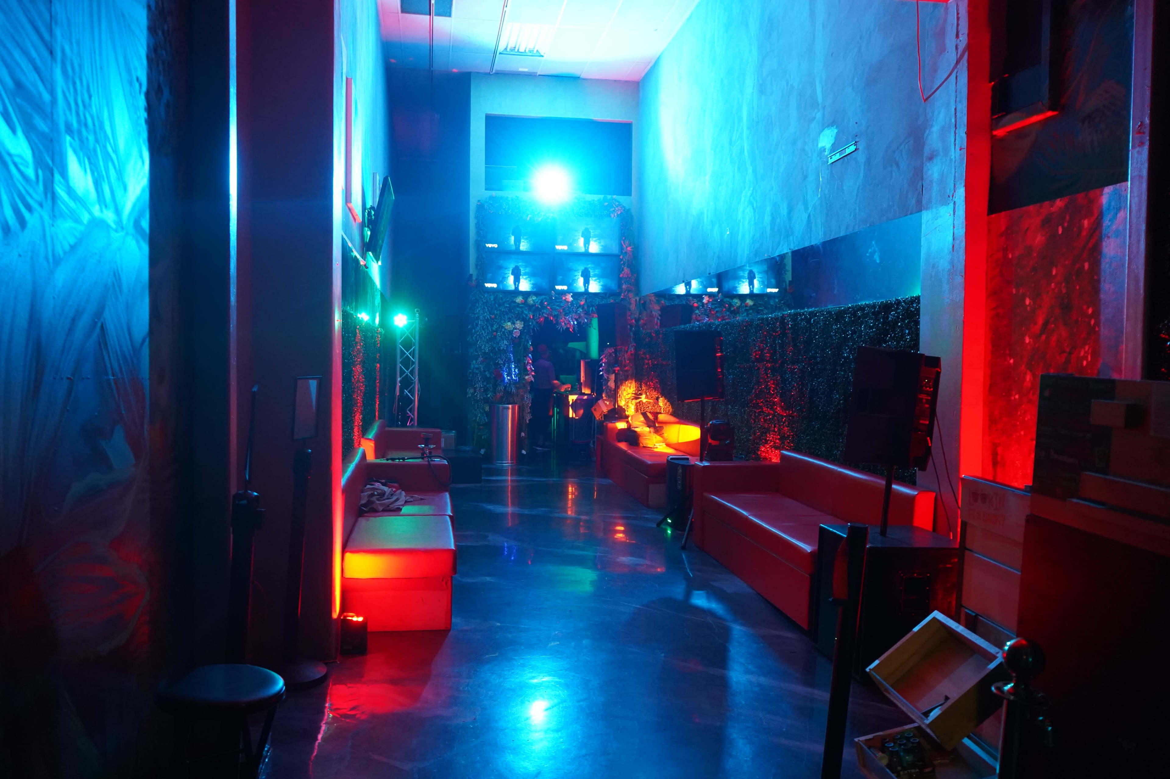 The image shows a dimly lit lounge area featuring red and blue lighting, with couches and screens lining the walls.
