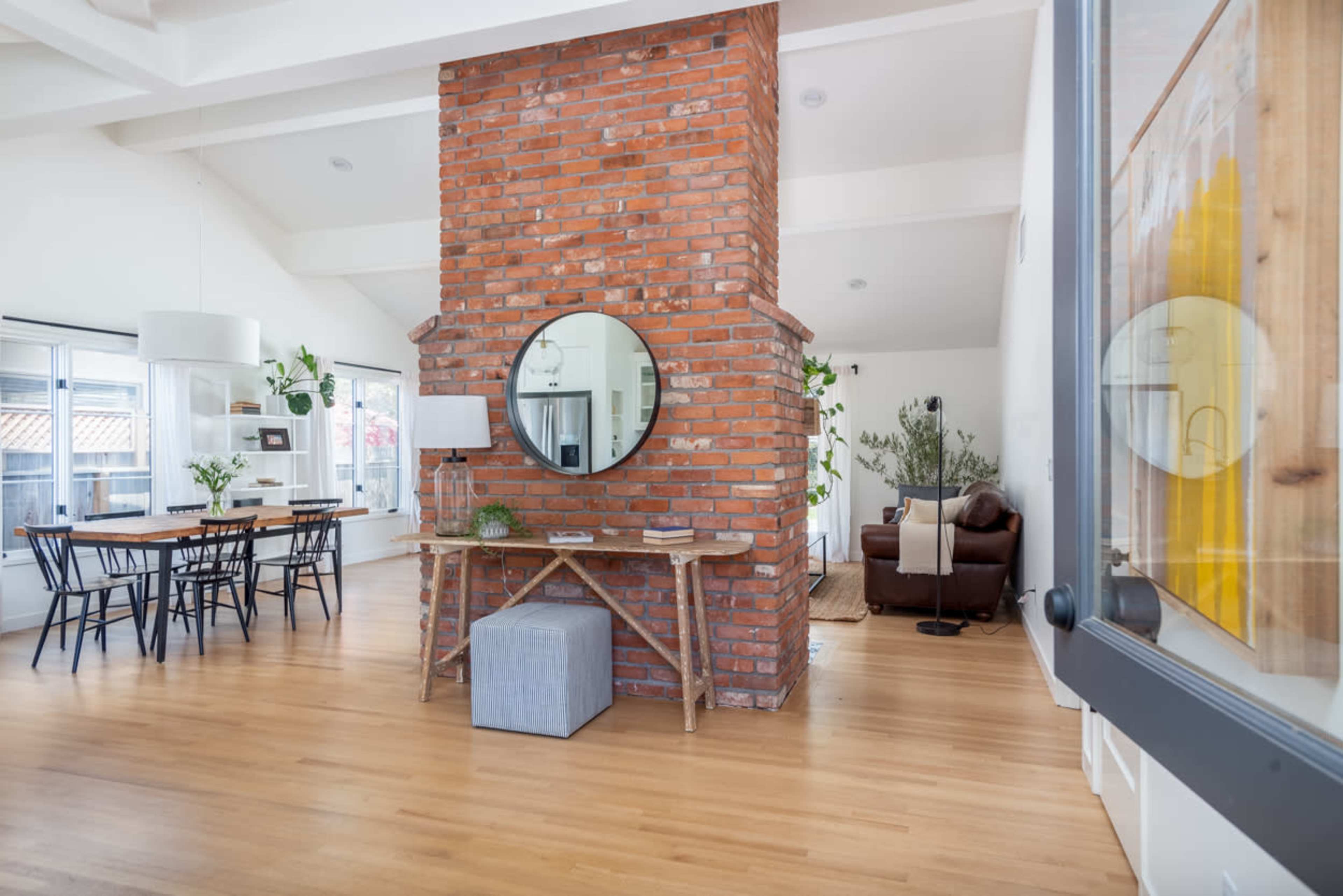 An open living space with a large brick column, a round mirror, a wooden dining table, and a cozy seating area.