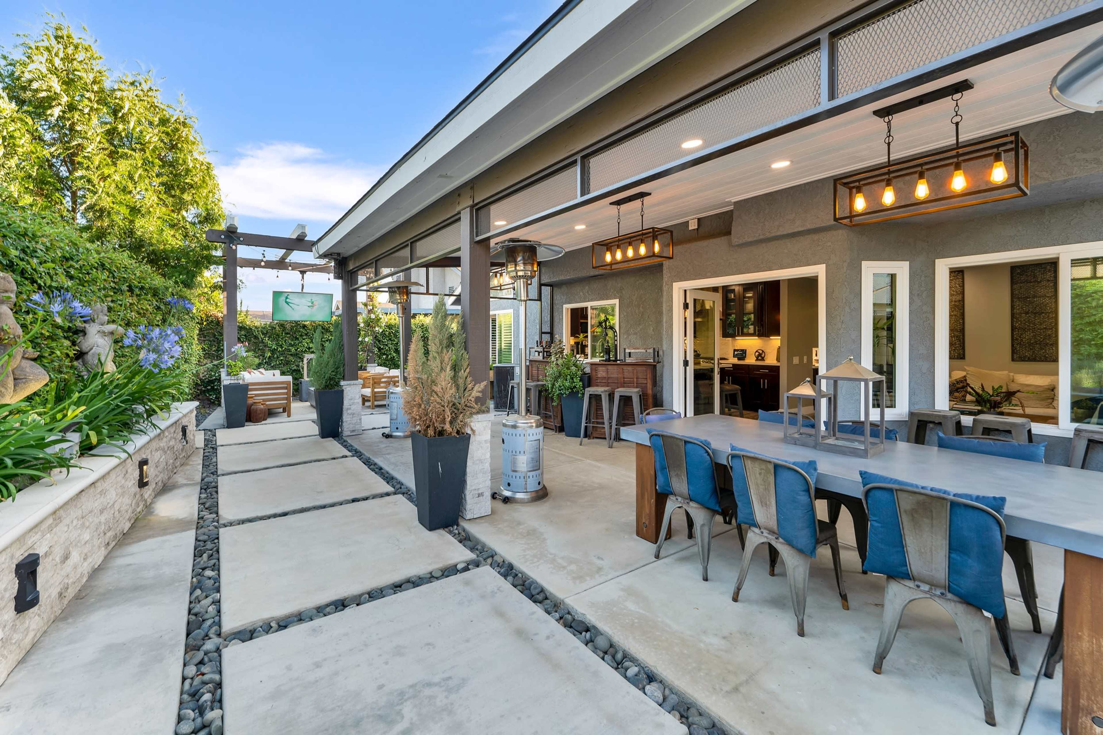 A modern outdoor patio area with a dining table, lounge seating, and a bar, surrounded by lush greenery and a stone pathway.