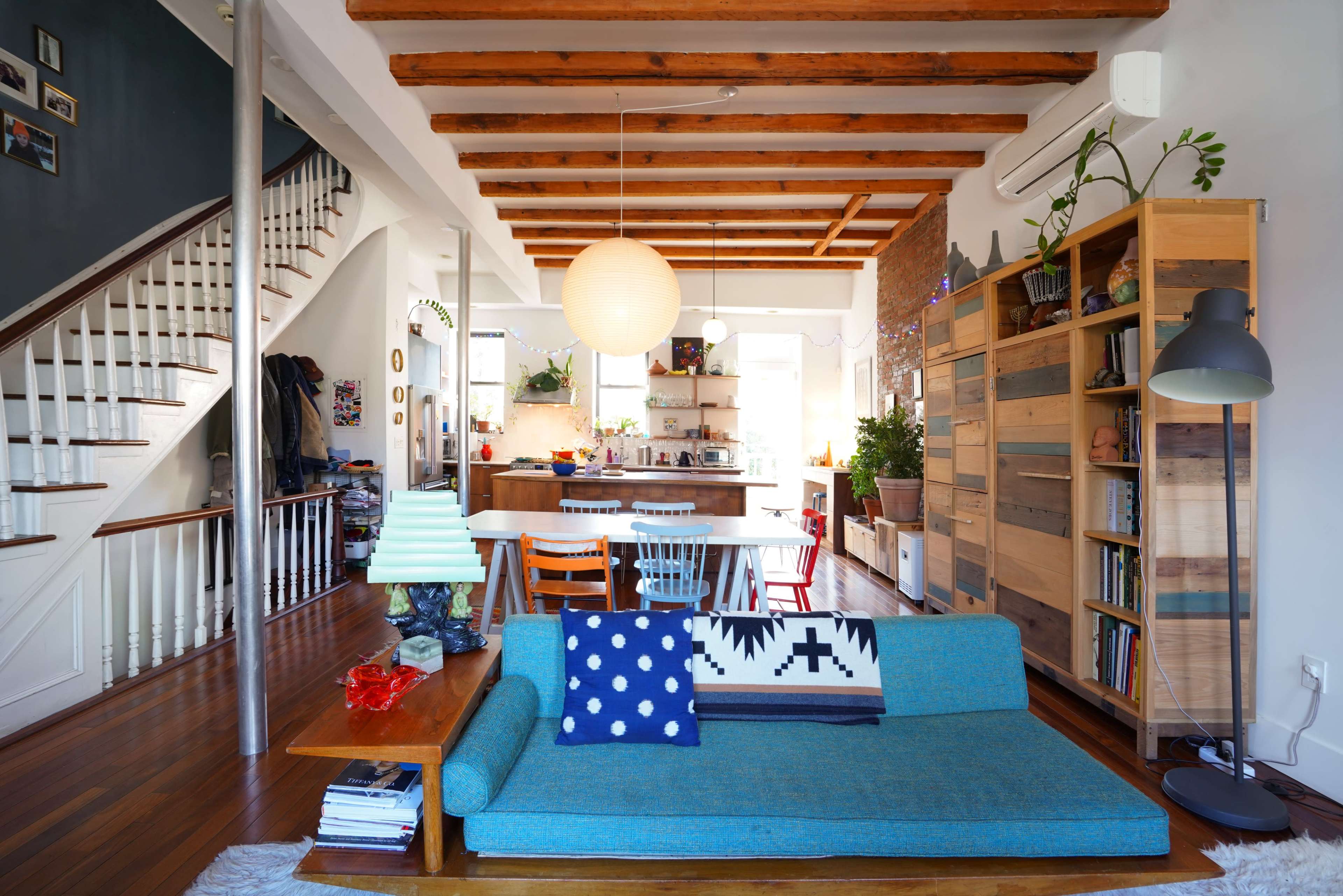 A modern living room with a blue sofa, wooden ceiling beams, and an open kitchen area visible in the background.