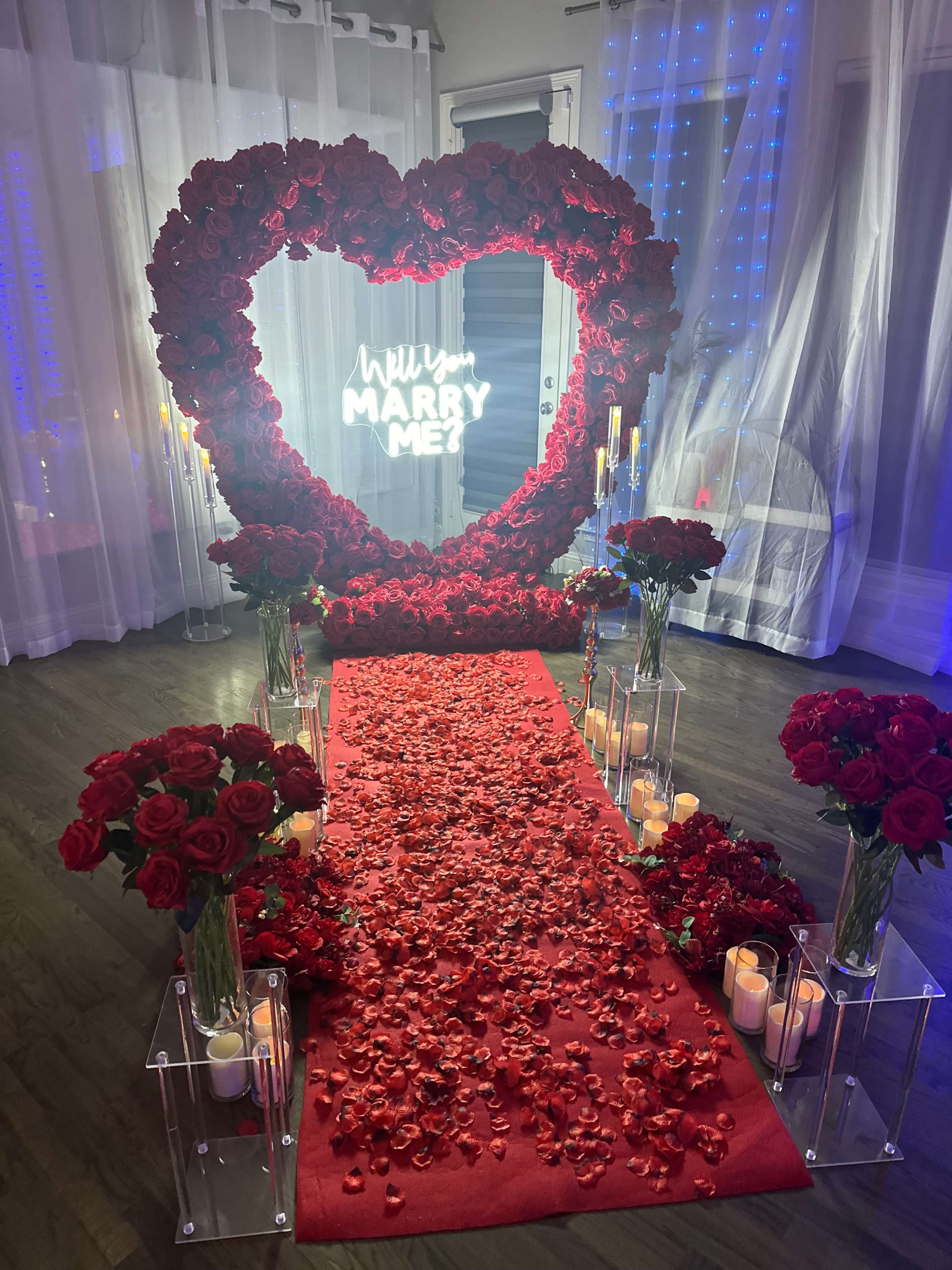 The image features a romantic setup for a marriage proposal, characterized by a heart-shaped flower arrangement, a red carpet, and candles surrounded by rose petals.