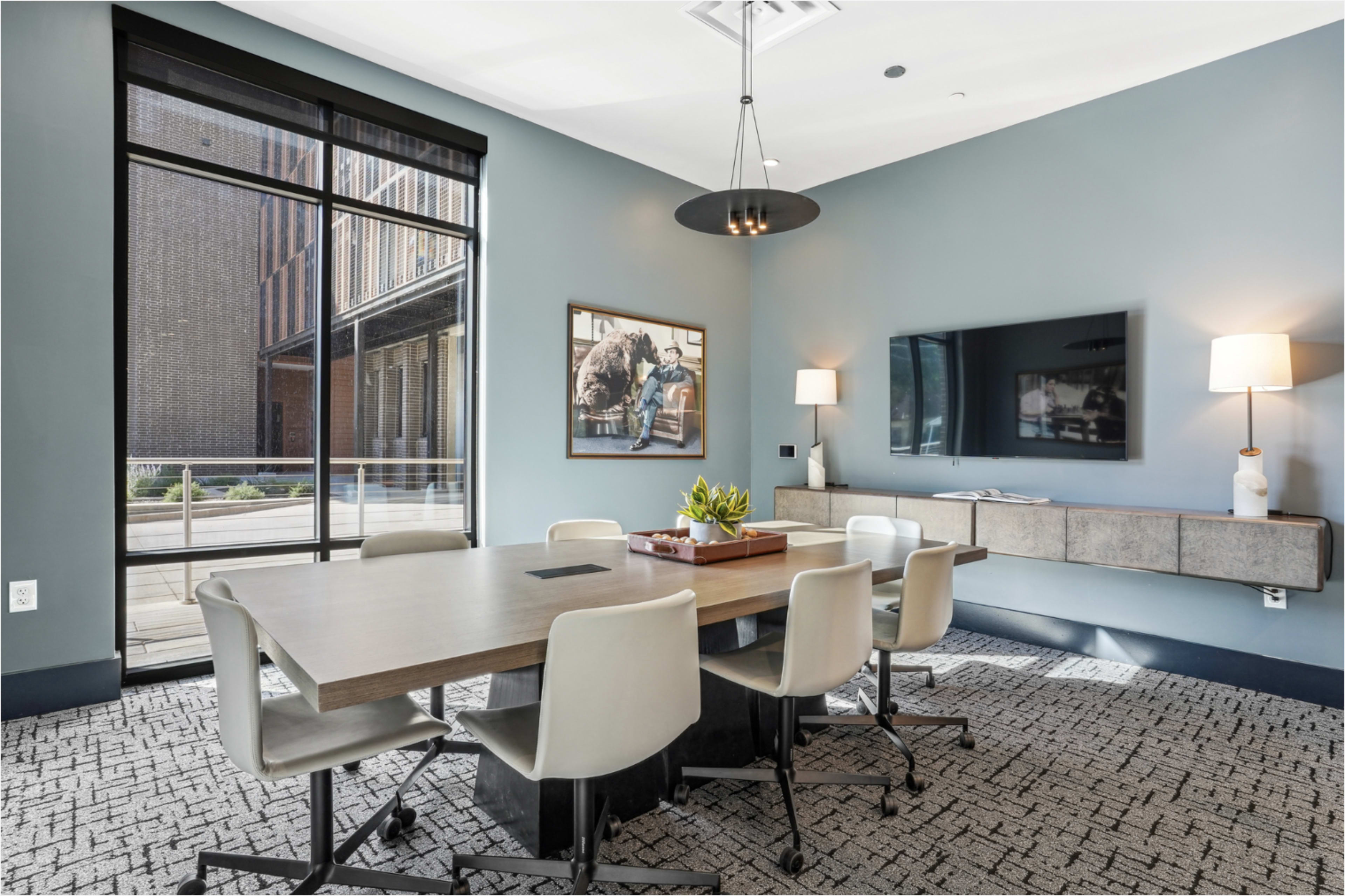 A modern conference room features a wooden table surrounded by ergonomic chairs, a television mounted on a wall, and a view of an outdoor area through large windows.