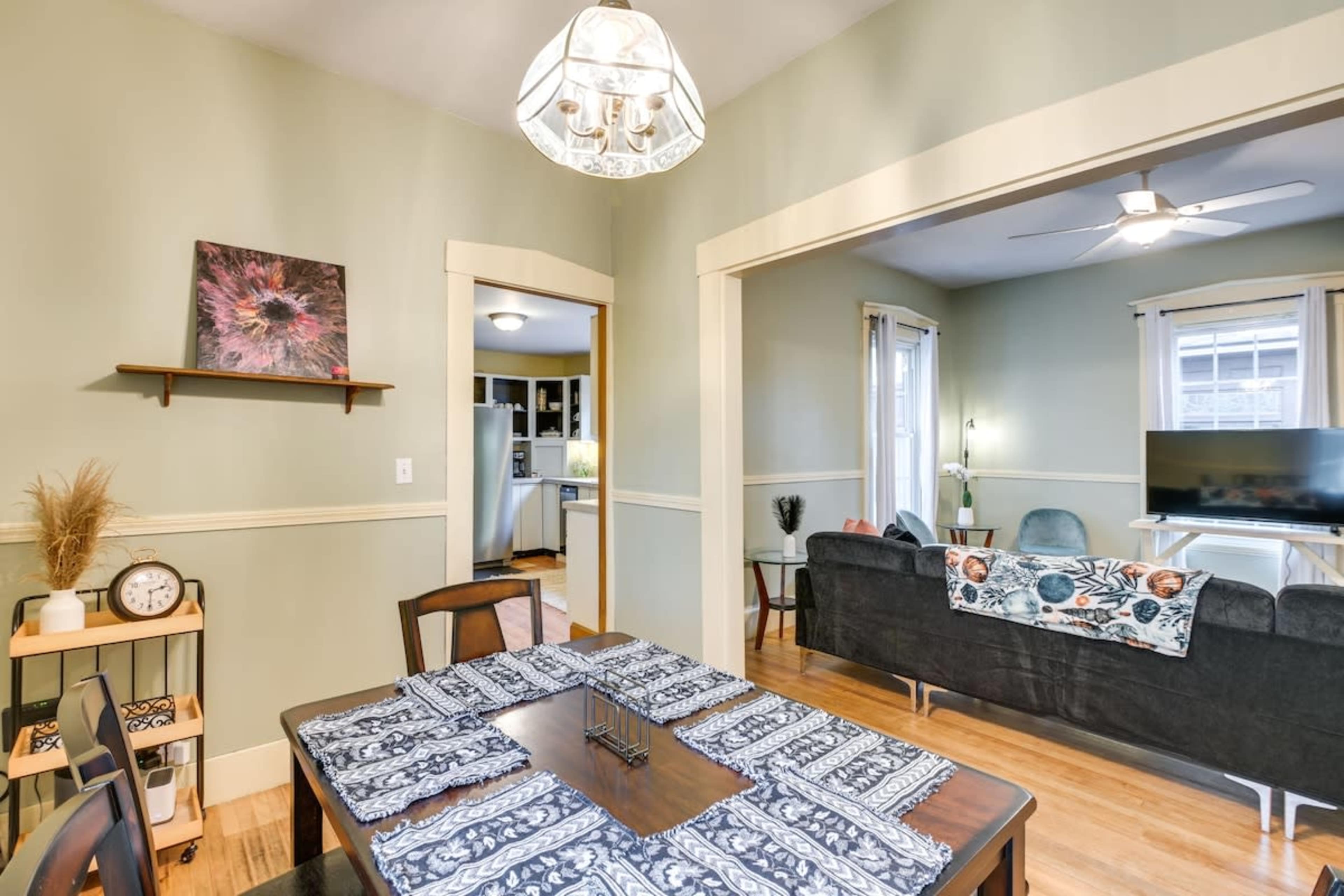 A dining area features a wooden table set with a patterned tablecloth, a clock, and a small shelf, leading into a living space with a couch and television.
