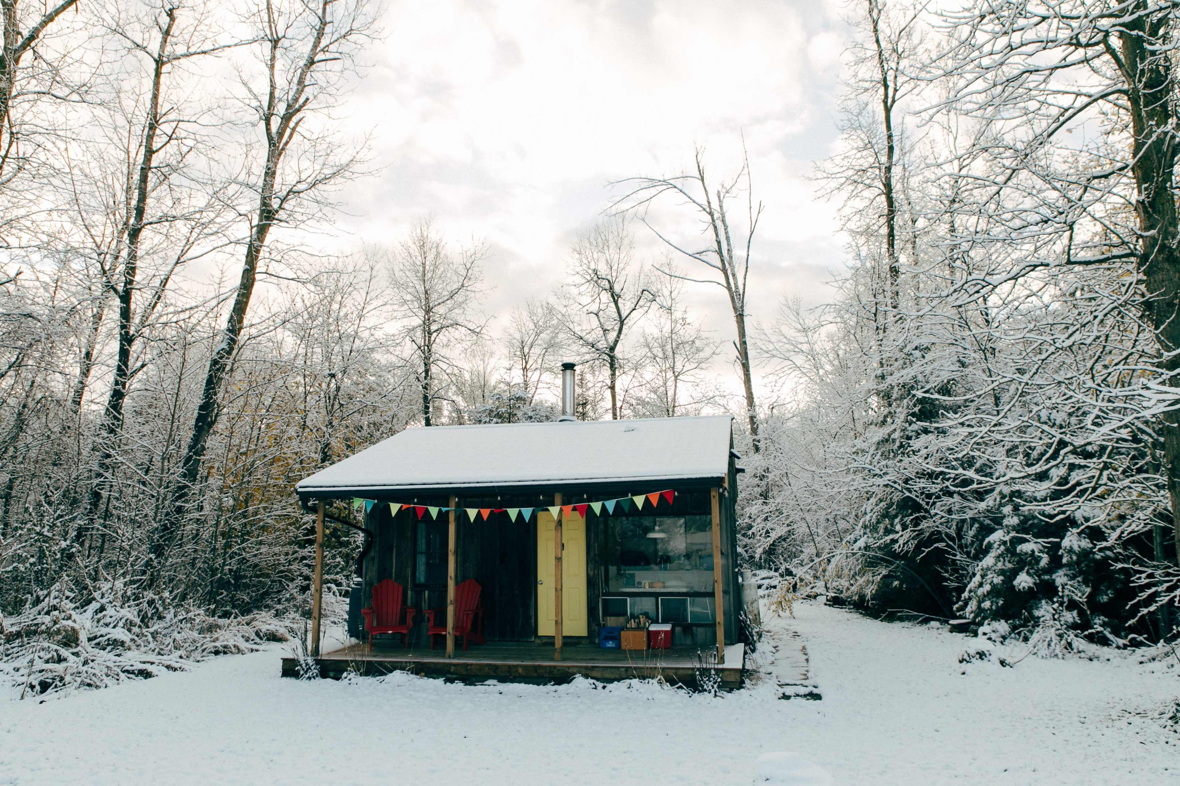 A small cabin is surrounded by snow-covered trees and has colorful bunting hanging on the porch.