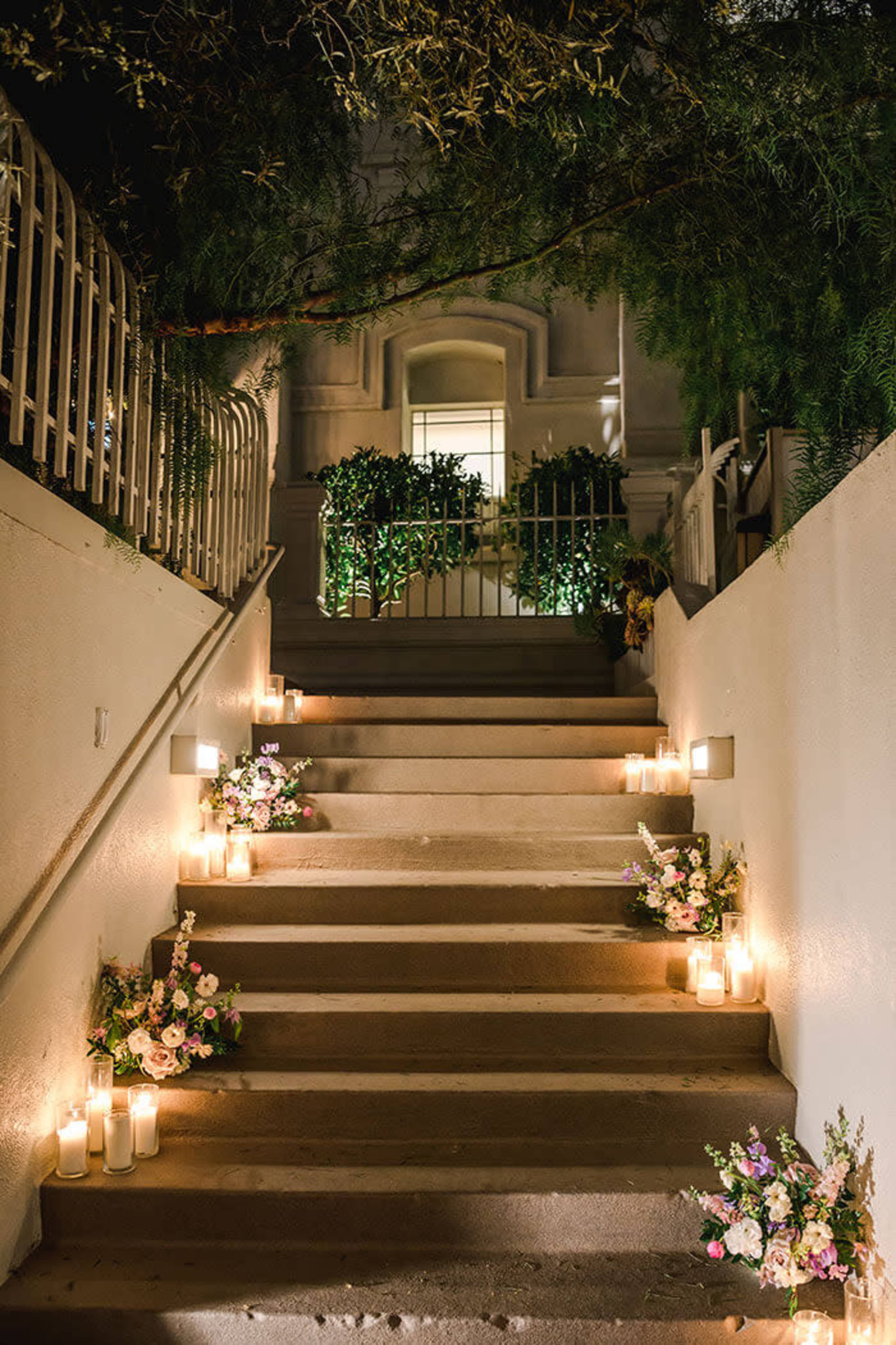 The image shows a staircase lined with glowing candles and floral arrangements on either side, leading up to a doorway surrounded by greenery.