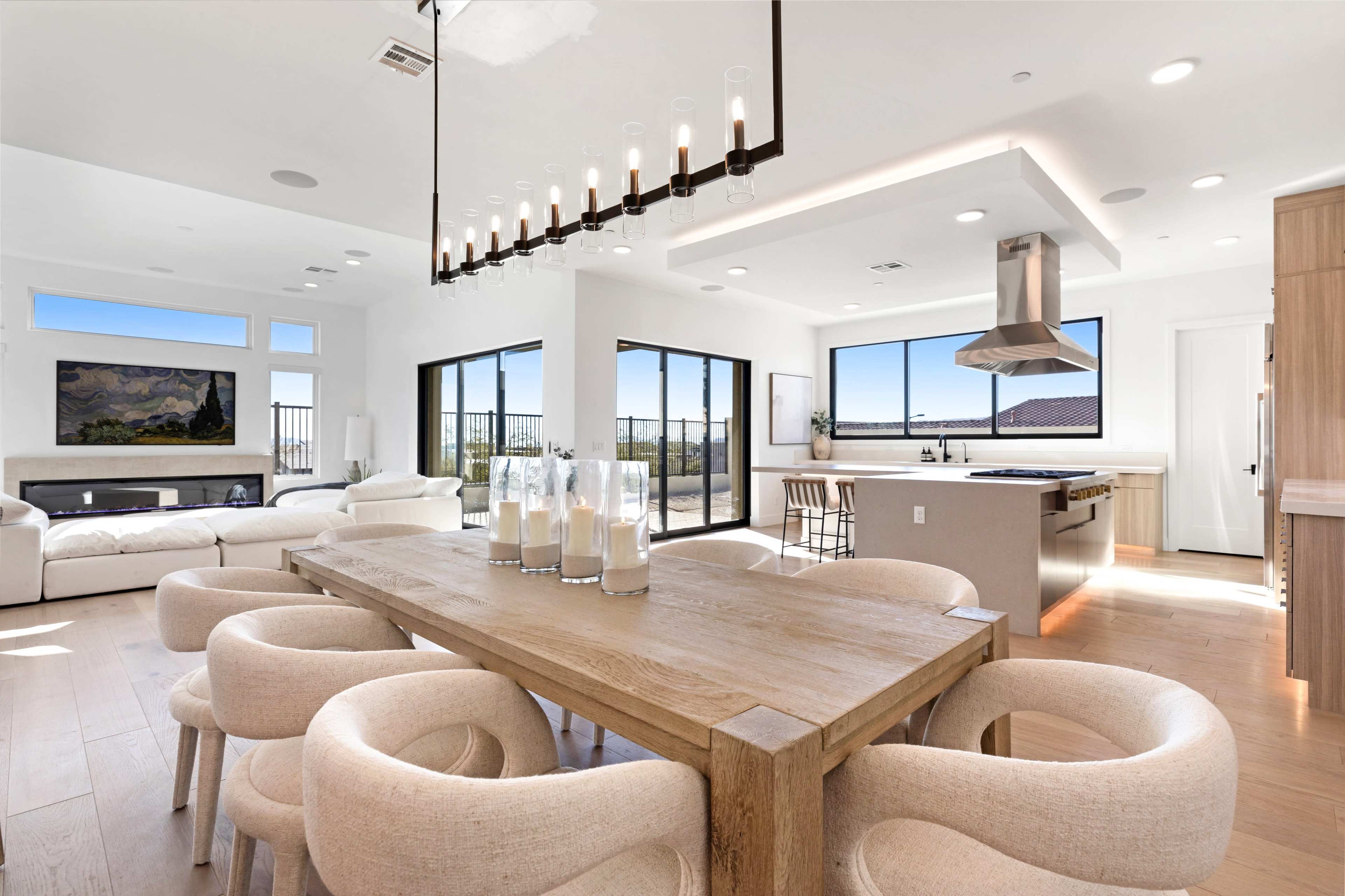 A modern open-concept living and dining area featuring a wooden dining table surrounded by upholstered chairs, a sleek kitchen with a central island, and large windows that provide natural light and outdoor views.