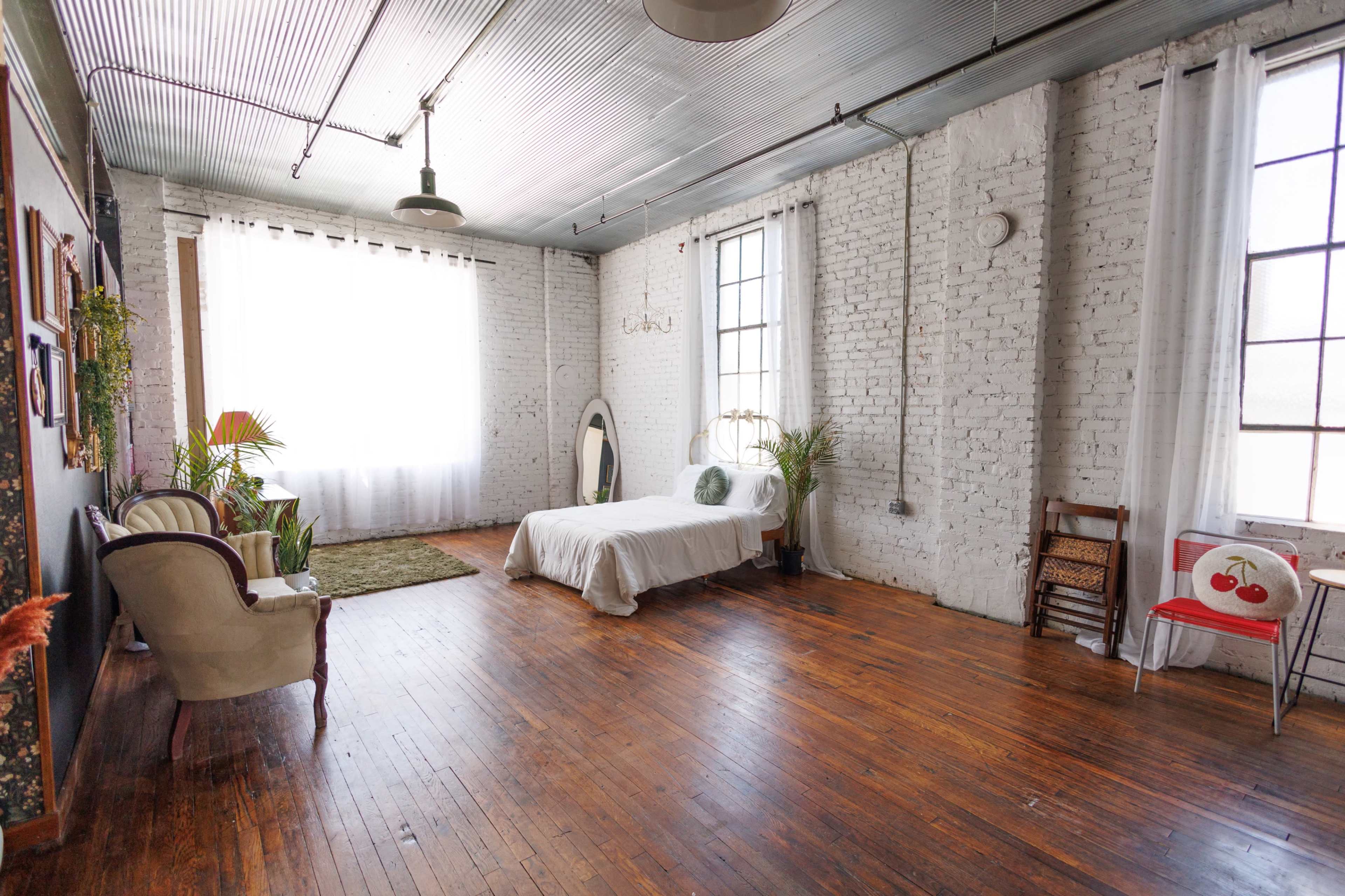 The image shows a spacious, well-lit room with white brick walls, large windows, wooden flooring, a bed, and various seating arrangements including a chair and a sofa.