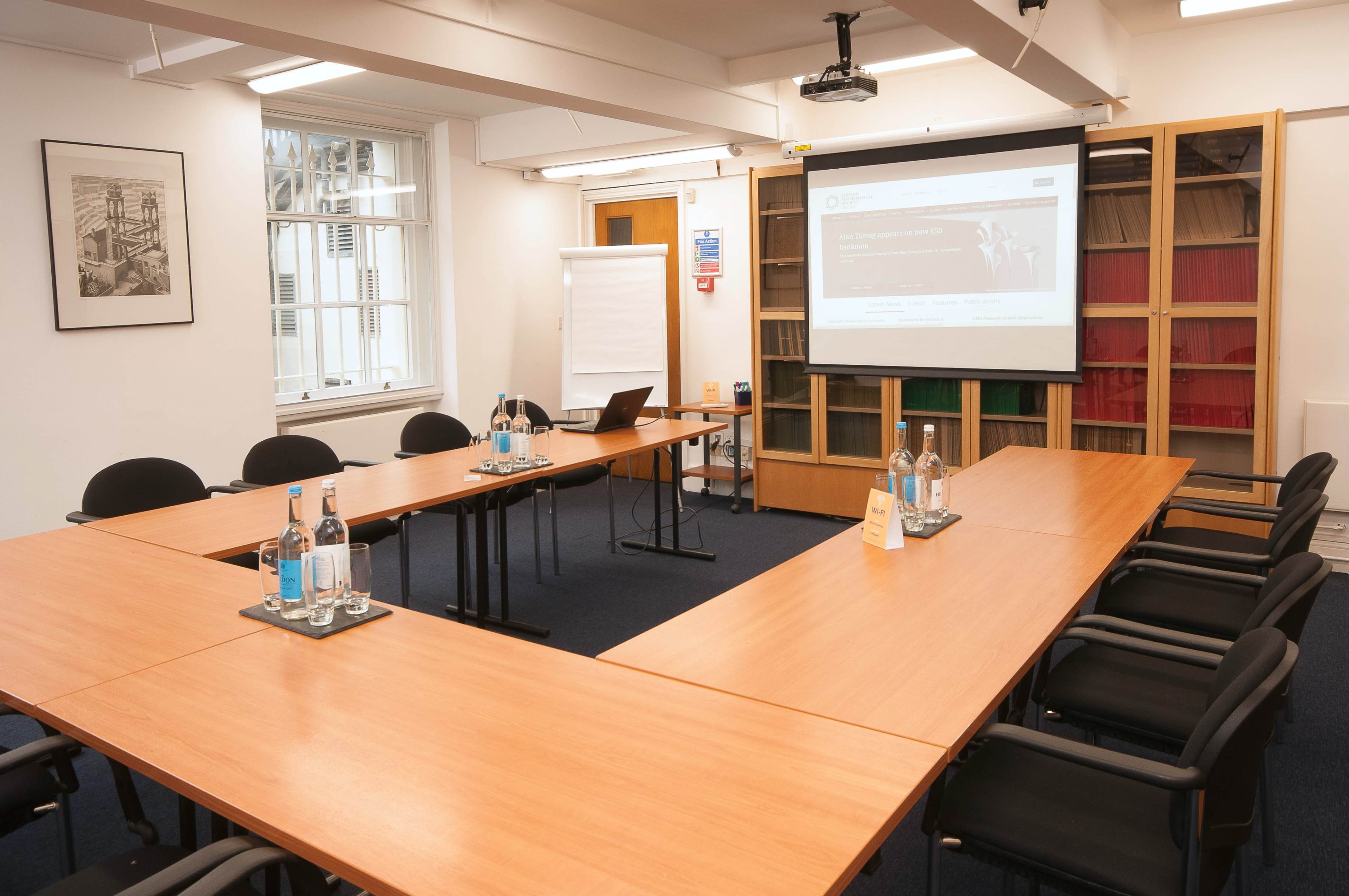 A conference room is arranged with a long, wooden table, black chairs, and a projector screen displaying a webpage, surrounded by shelves containing books.