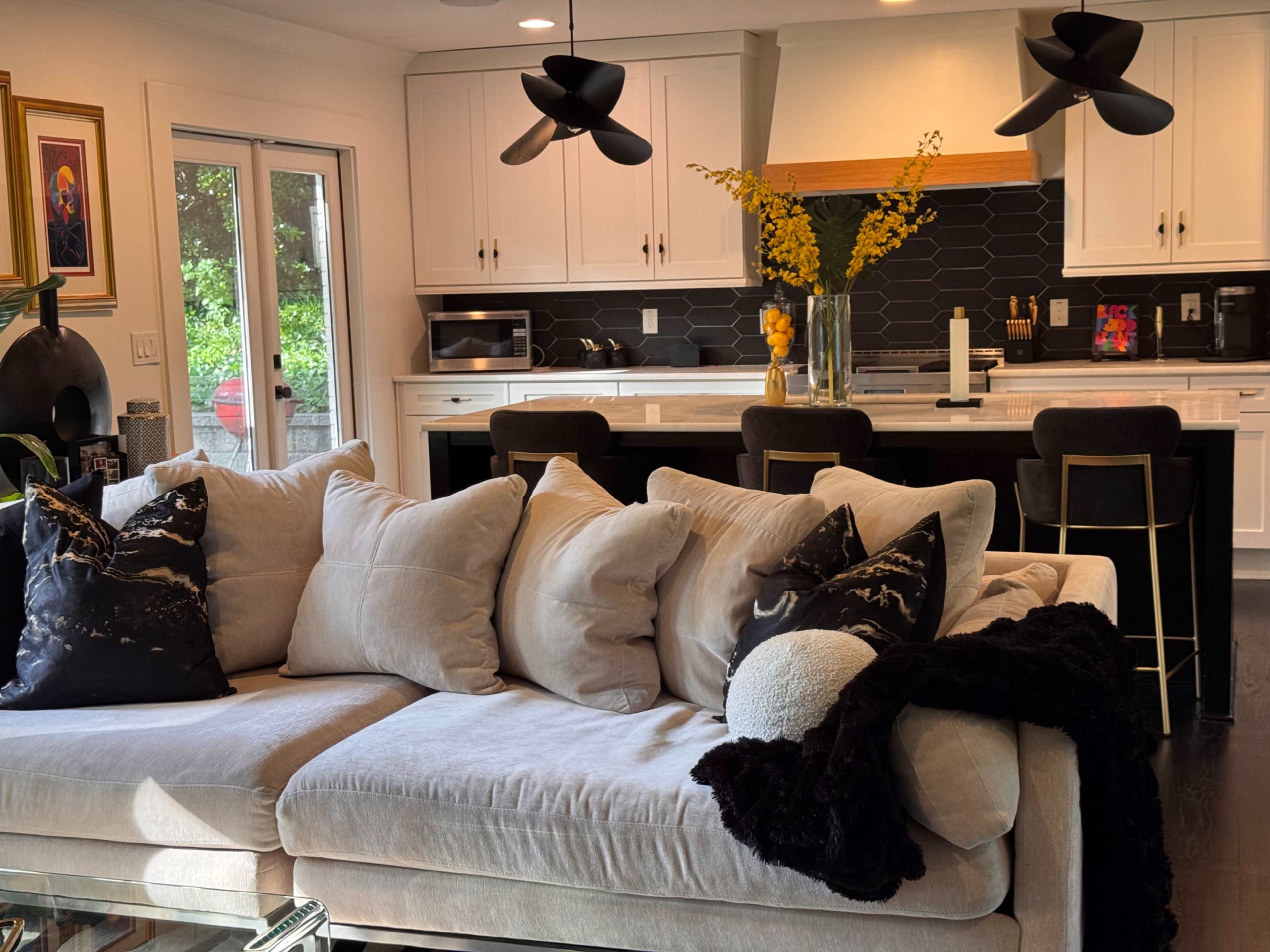 A light-colored sectional sofa with multiple decorative pillows is positioned in a modern kitchen and living area featuring dark countertops and a dining bar.