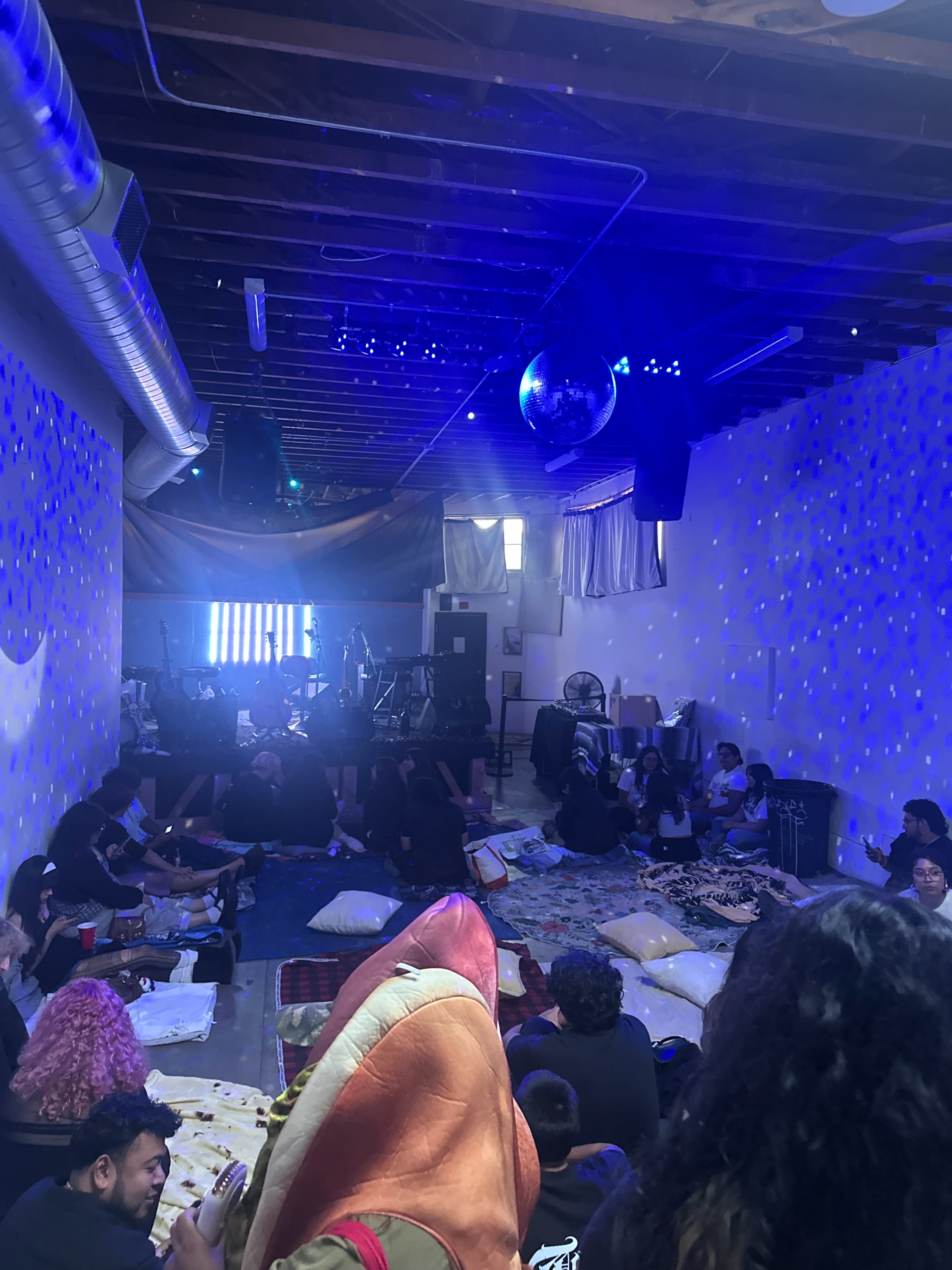 A group of people is sitting on the floor of a dimly lit room with colorful lighting and musical equipment set up at the front.