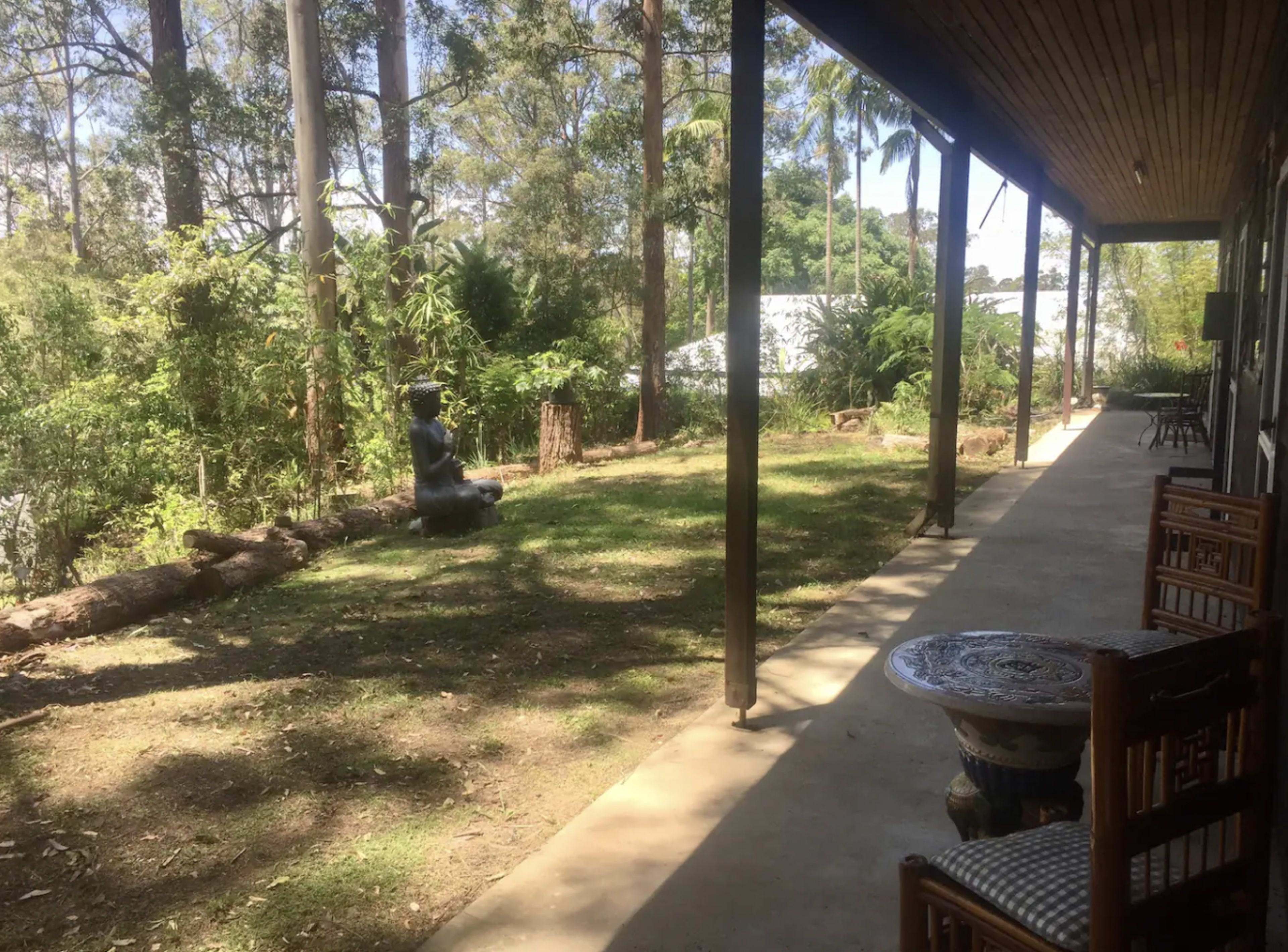 A porch extends along a house, overlooking a grassy area with a seated statue surrounded by trees and shrubs.