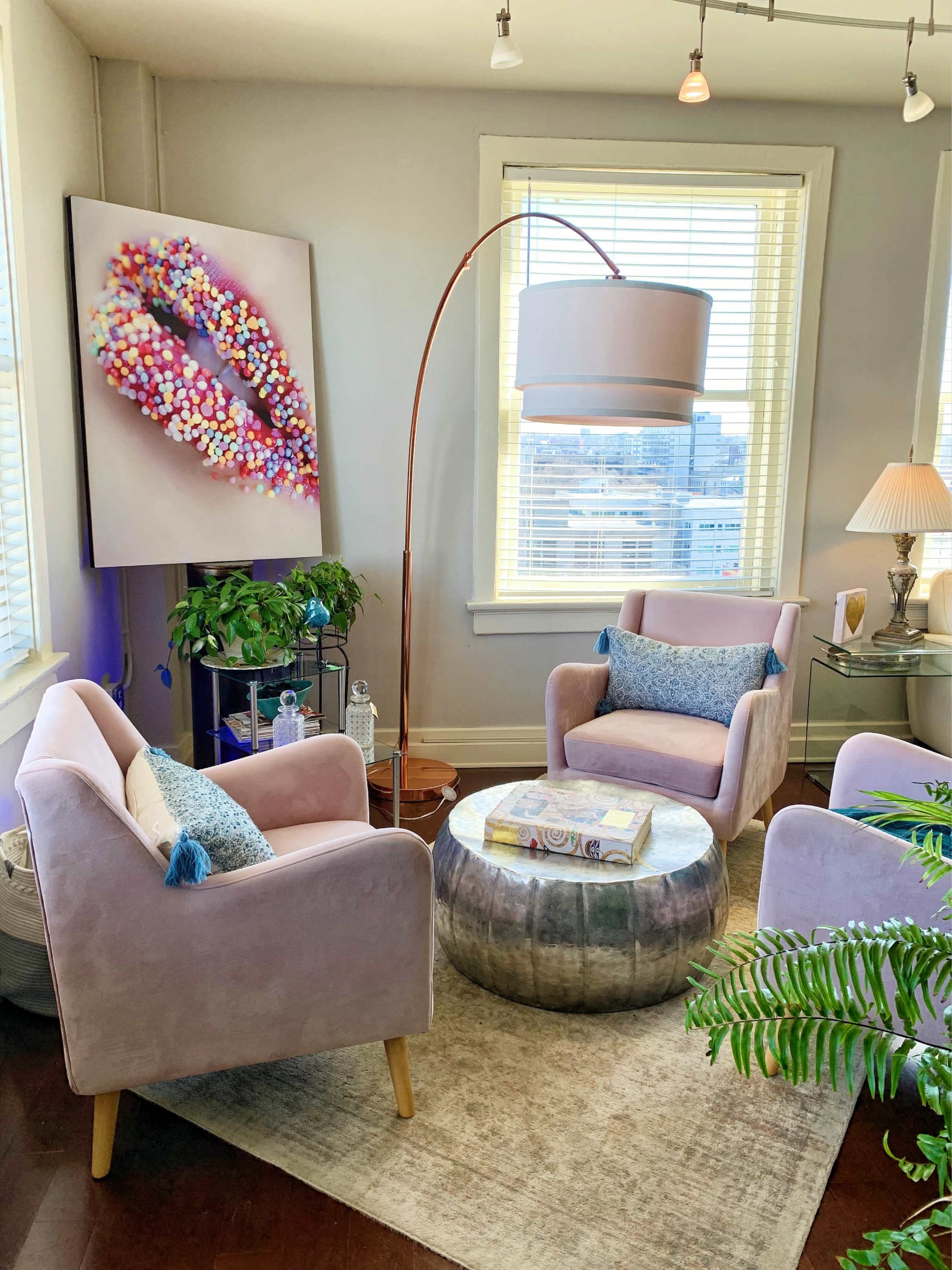 A cozy seating area with two pink chairs, a round metallic coffee table, a floor lamp, and a large colorful artwork on the wall.