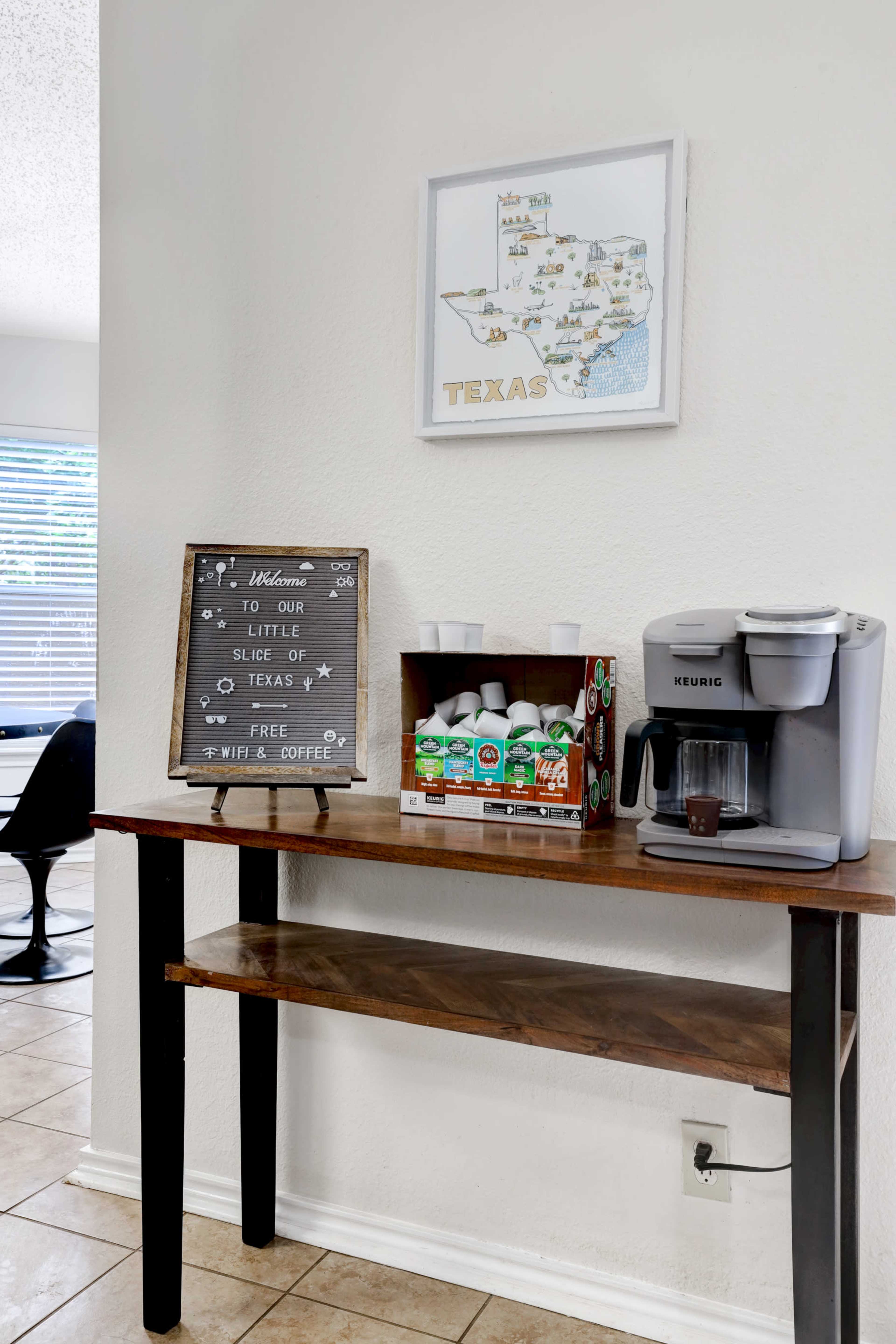 A wooden table displays a Keurig coffee machine, a box of creamers, and a welcome sign near a map of Texas.