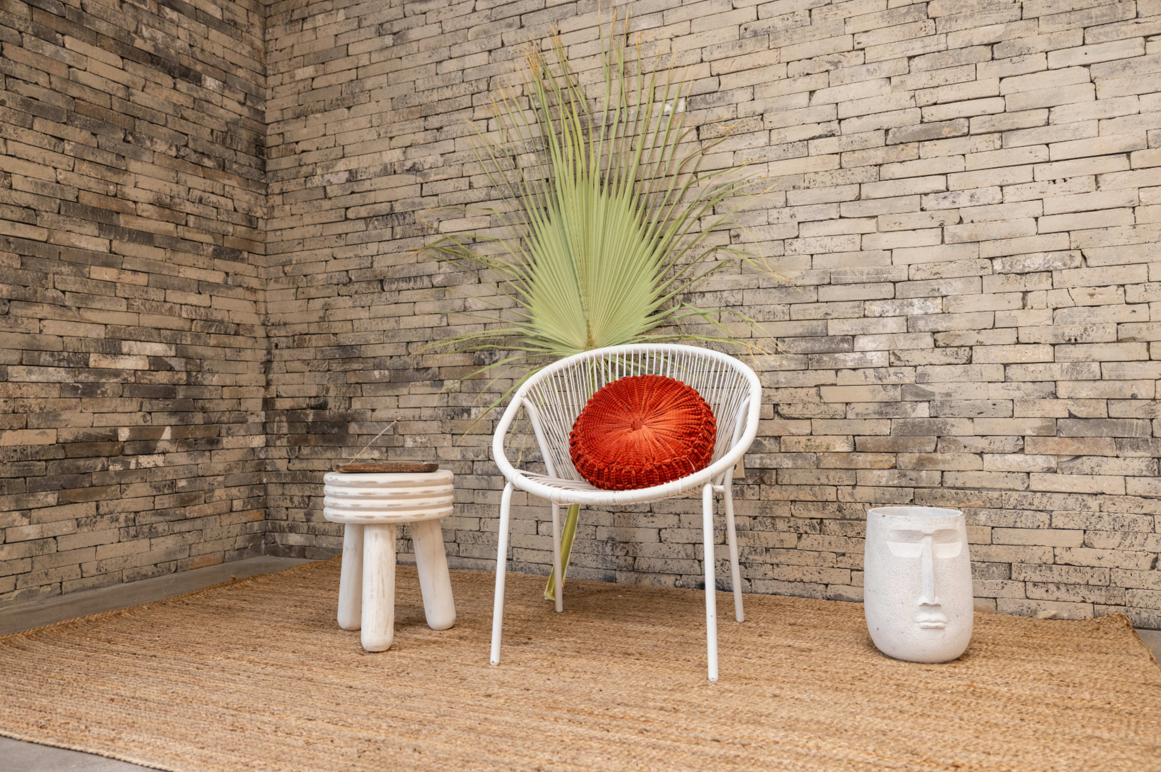 A modern seating area features a white chair with a round red cushion, a textured rug, a wooden stool, and a decorative stone face, all against a brick wall with a large green plant.