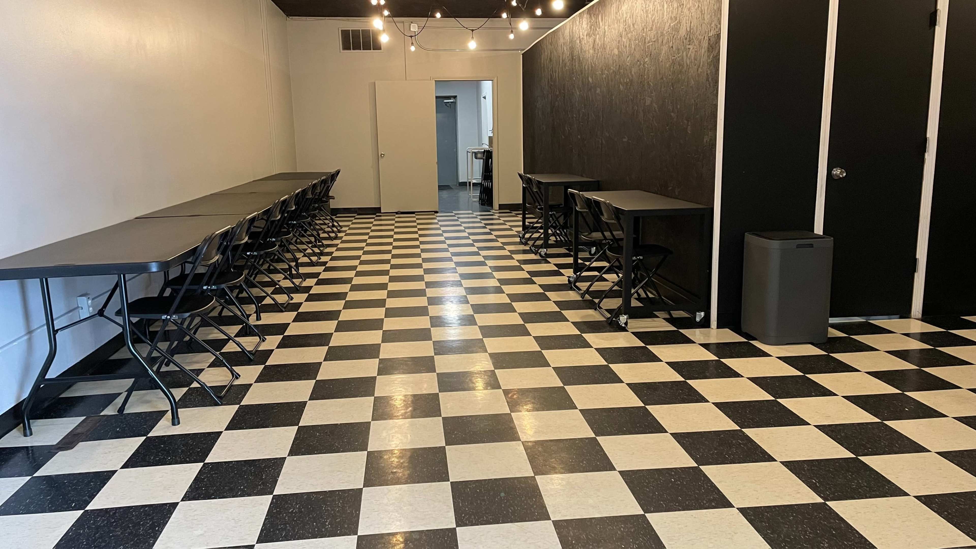 A room with a checkered black-and-white floor, featuring several tables and chairs lined along the walls, and a couple of doors at the far end.