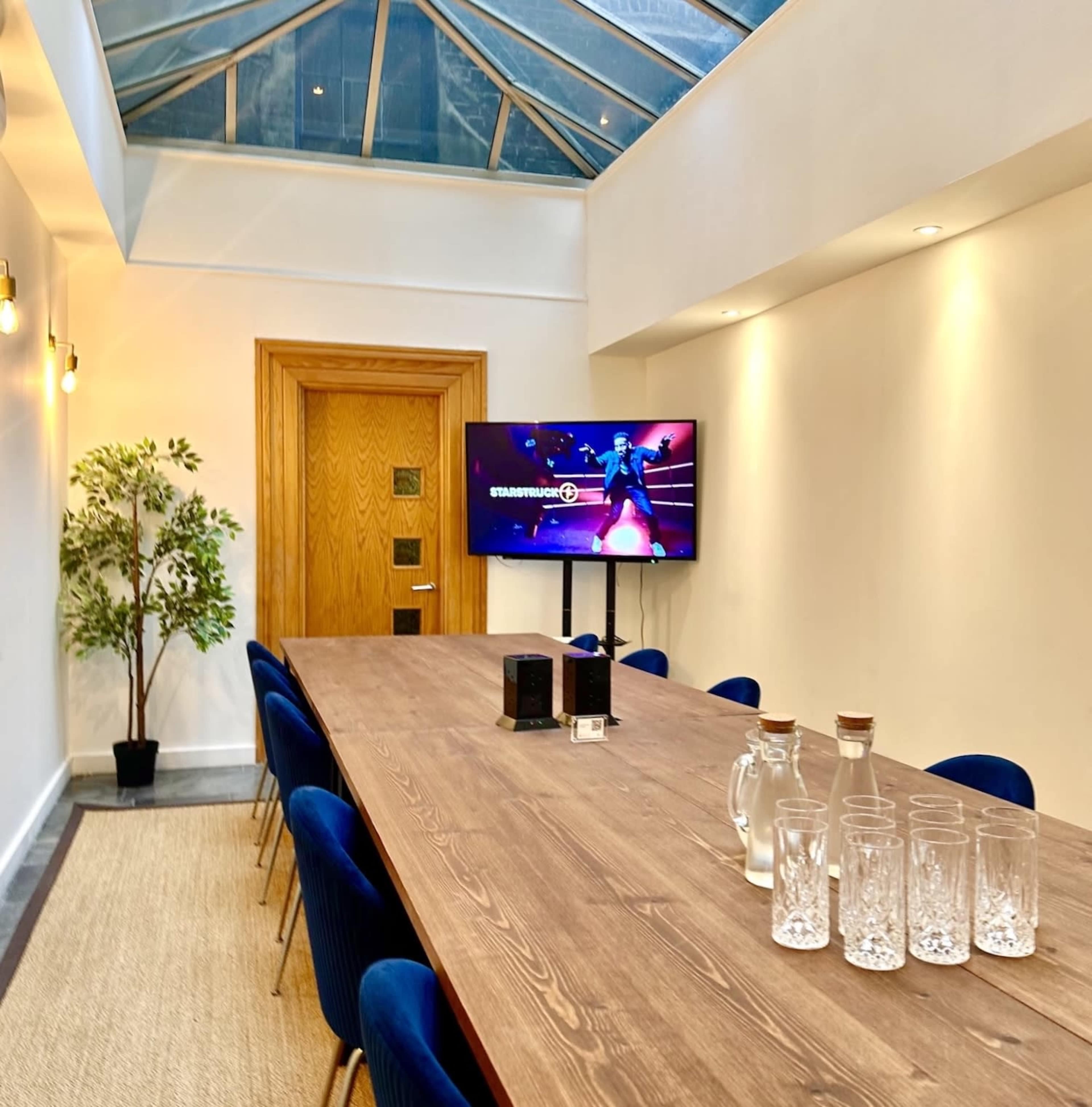 A long wooden conference table with blue chairs is set up in a well-lit meeting room featuring a large television screen on the wall and a potted plant in the corner.