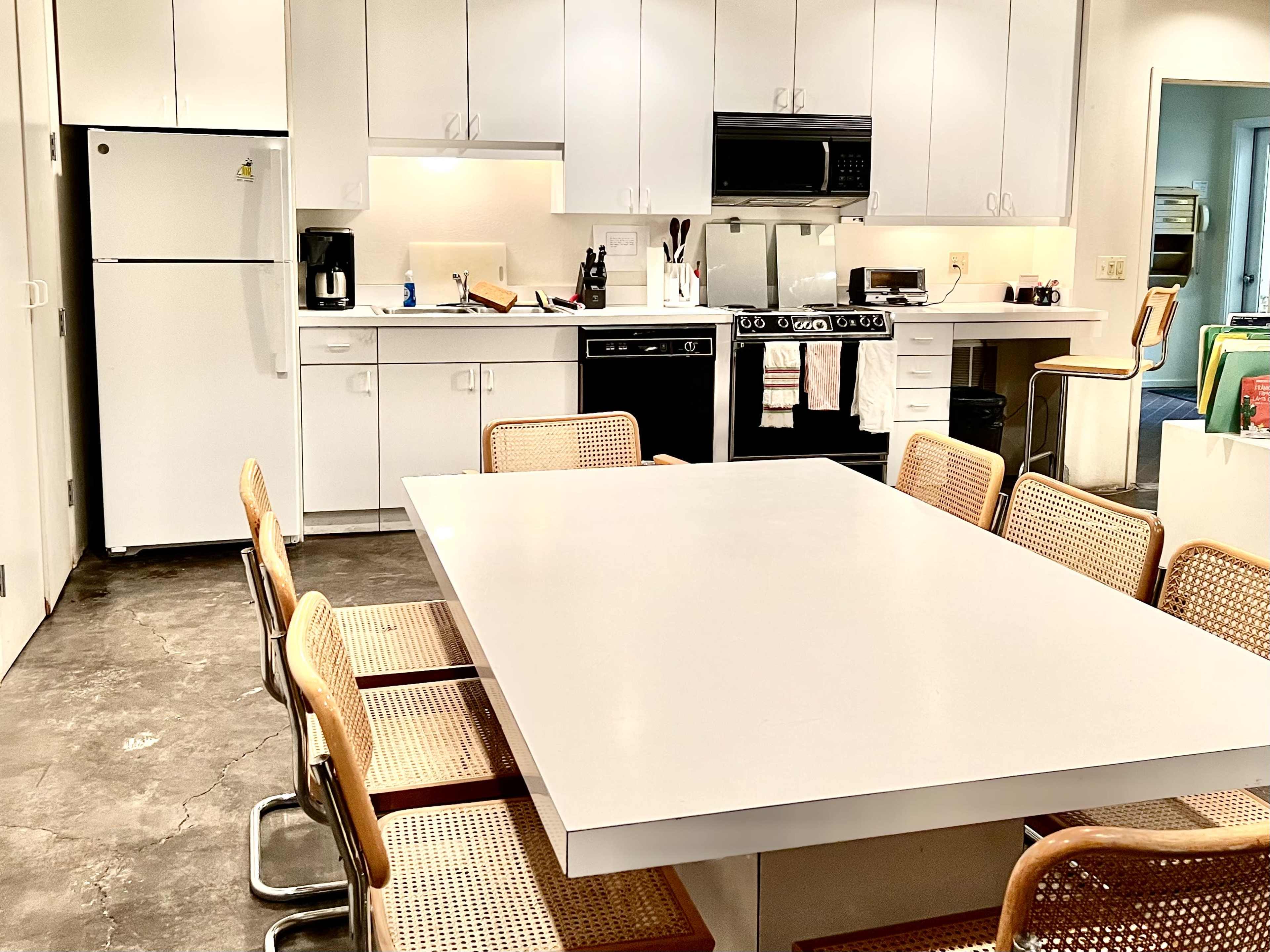 A modern kitchen features a large white table surrounded by eight cane-seat chairs, with appliances and cabinets in the background.