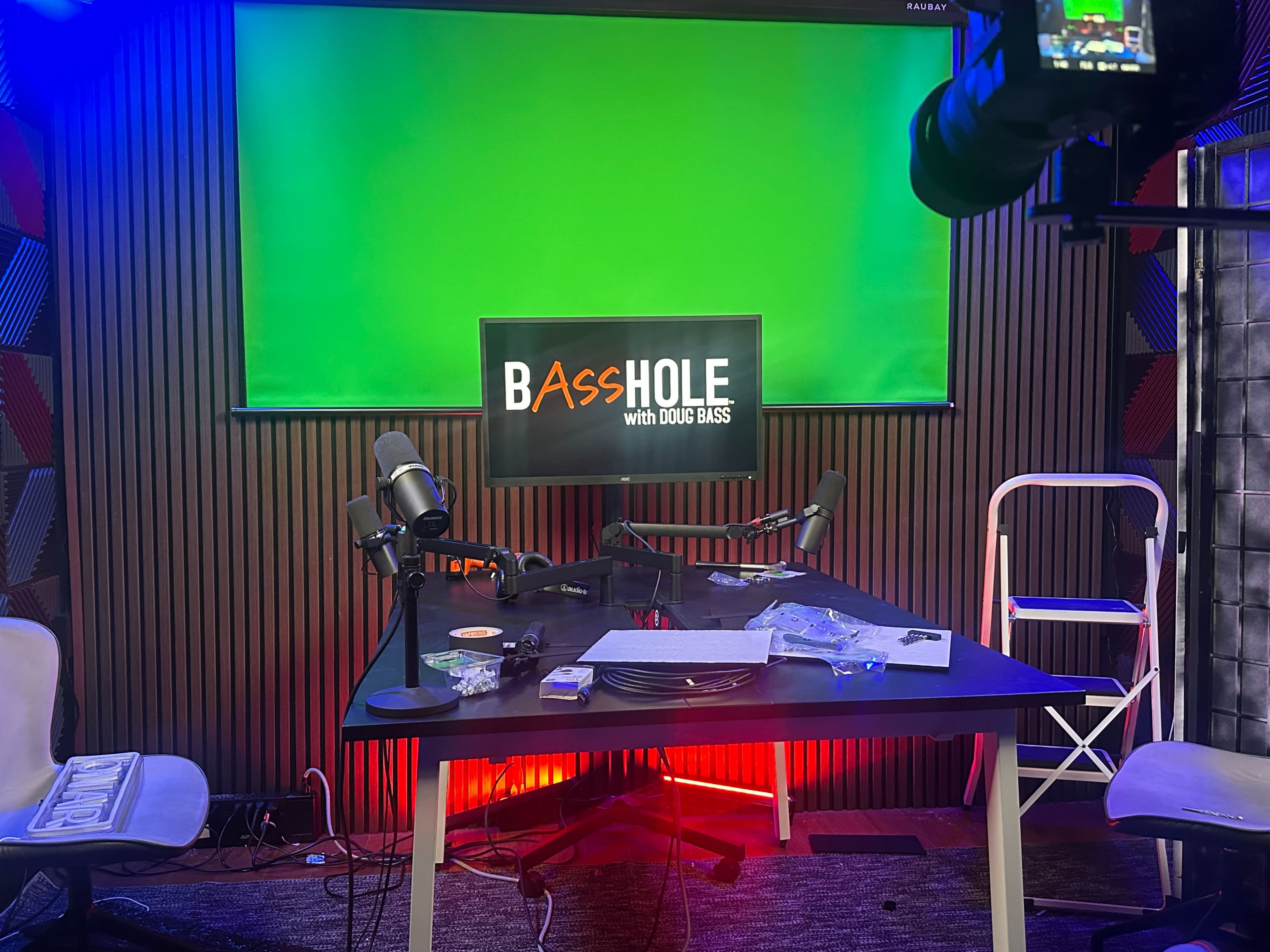 The image shows a podcast studio setup with a green screen backdrop, a table equipped with microphones, and a monitor displaying the title "BASSHOLE with Doug Bass."