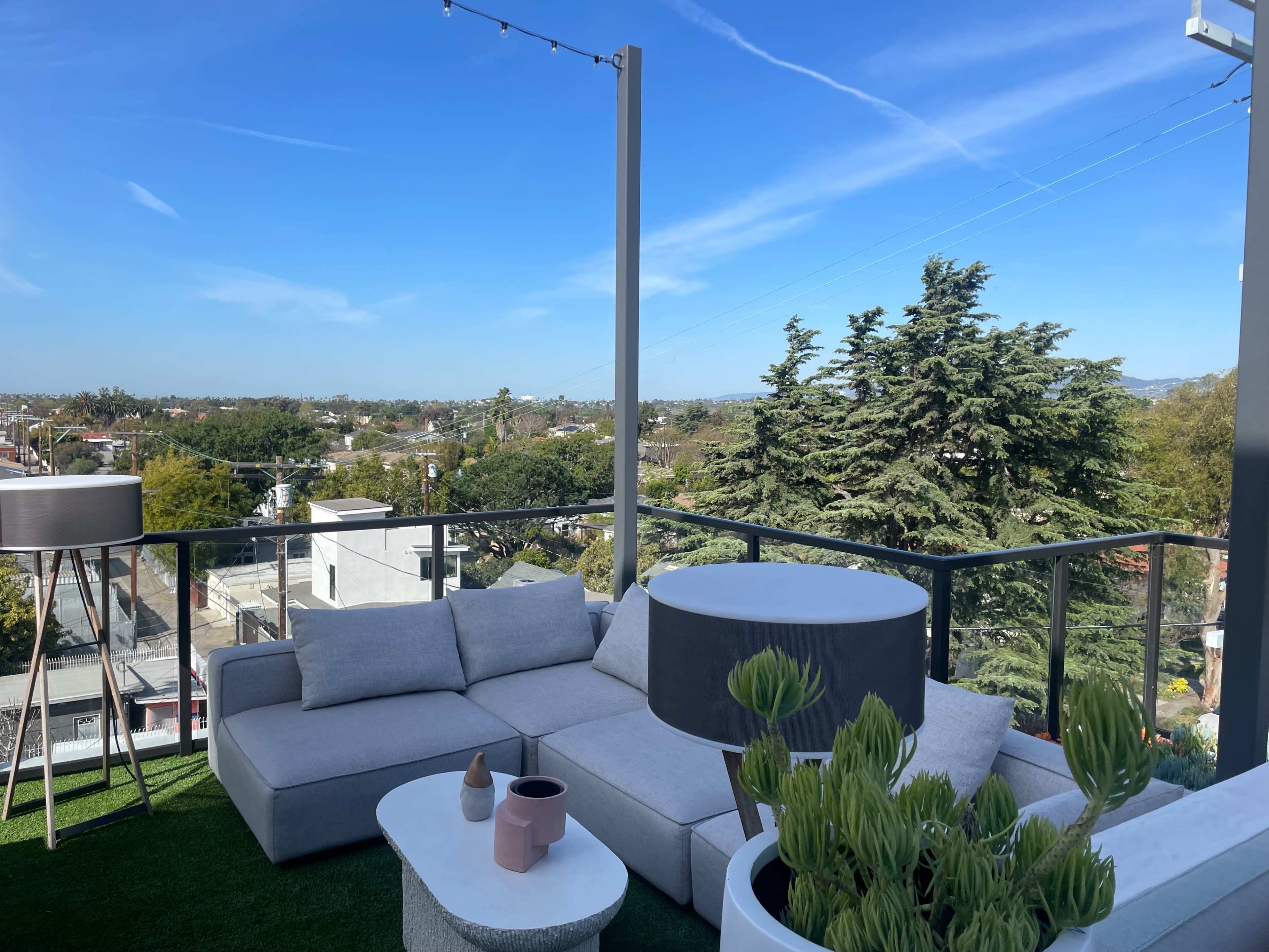 A modern outdoor seating area on a balcony, featuring a sectional sofa, a round table, and potted plants, with a view of a suburban landscape and clear blue sky.