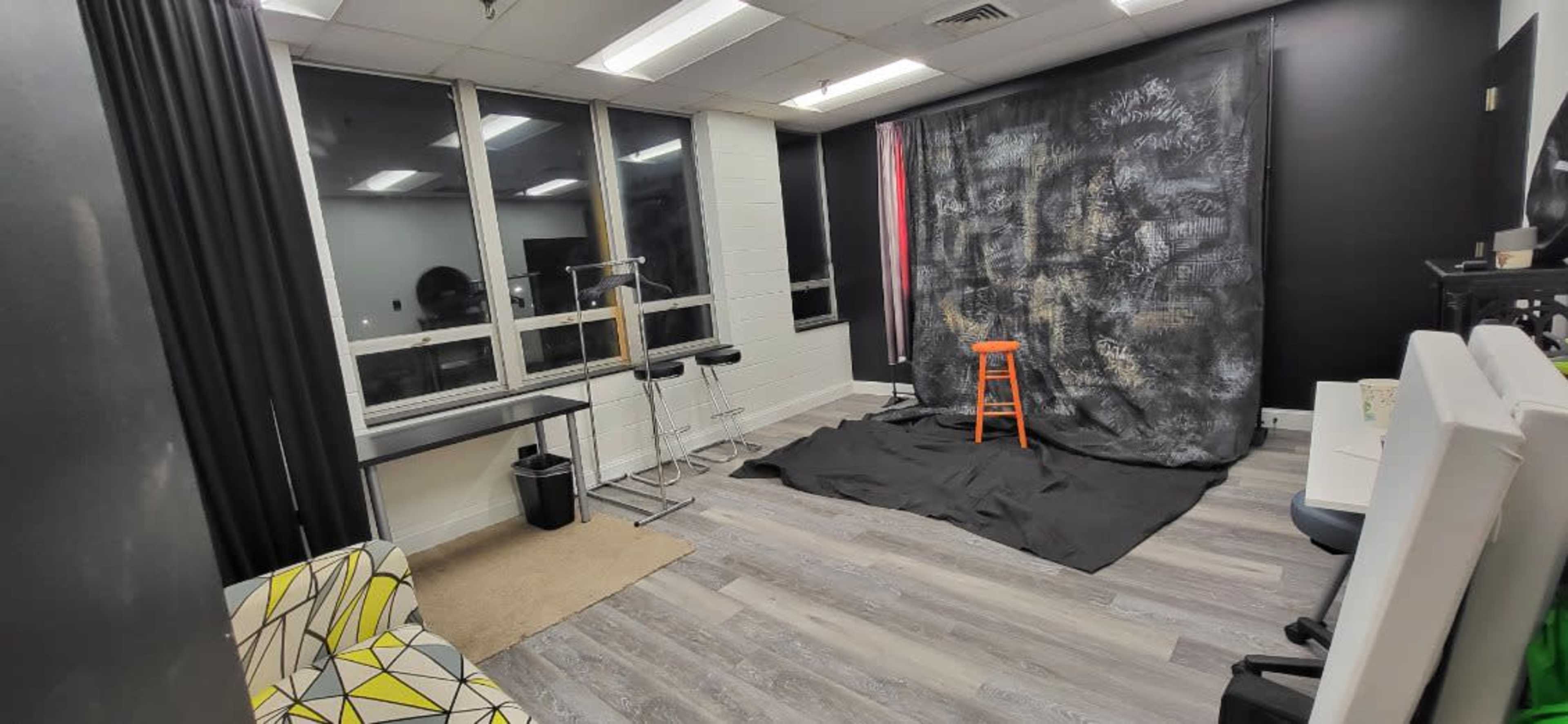 The image shows a sparsely furnished studio space featuring a black backdrop with a stool in front, a table on the side, and windows allowing natural light.