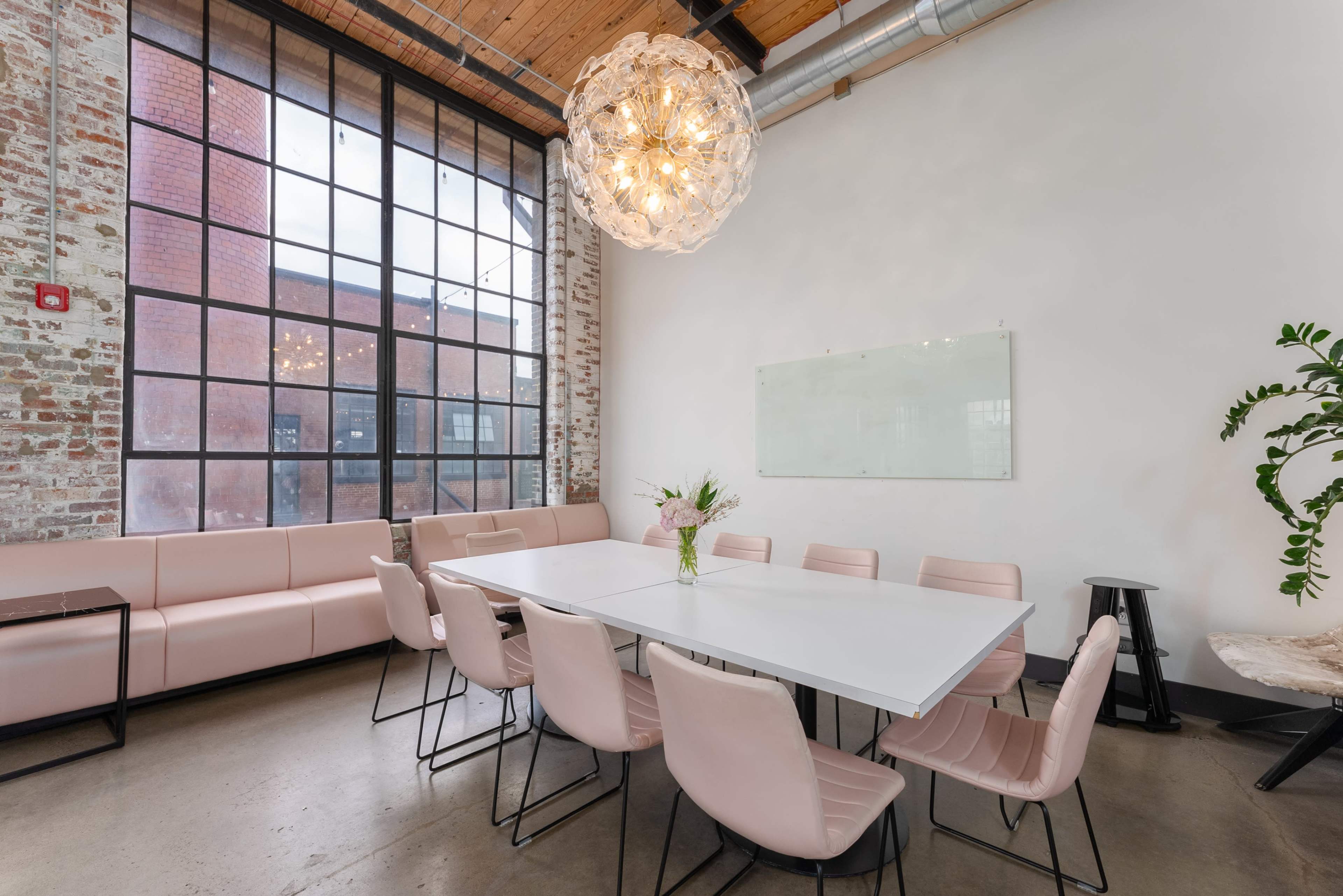 A modern meeting room features a long white table surrounded by pink chairs, with large windows and exposed brick walls.