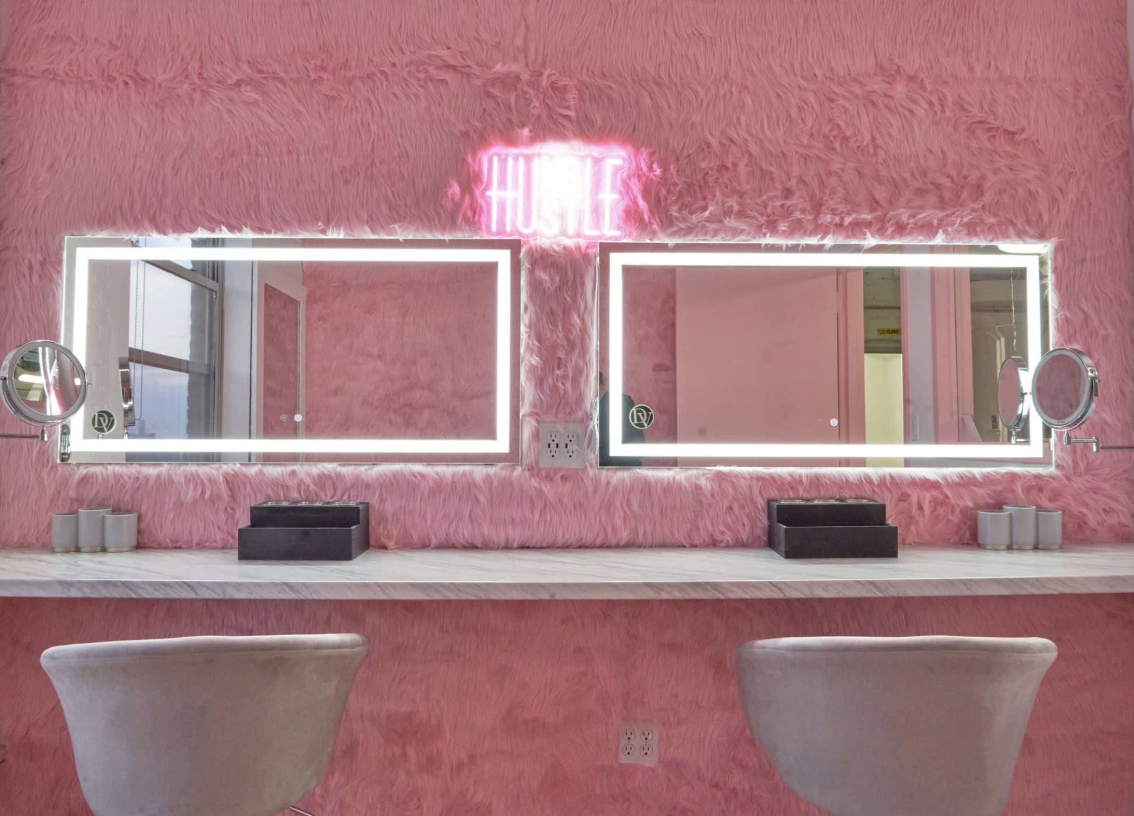 A vanity area features two illuminated mirrors set against a plush pink wall, with a neon sign that says "HUSTLE" above them.