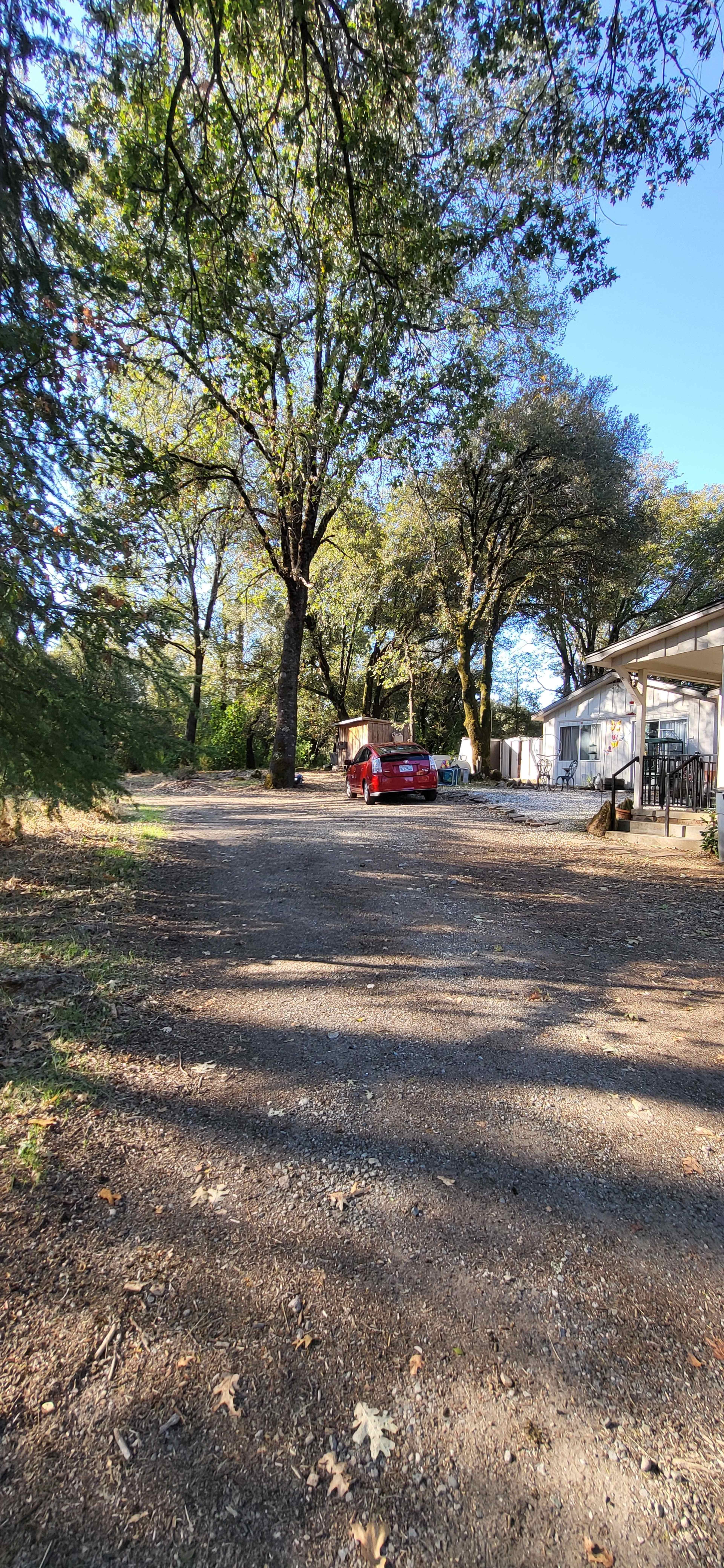 A red car is parked on a gravel driveway lined with trees, leading to a building in the background.