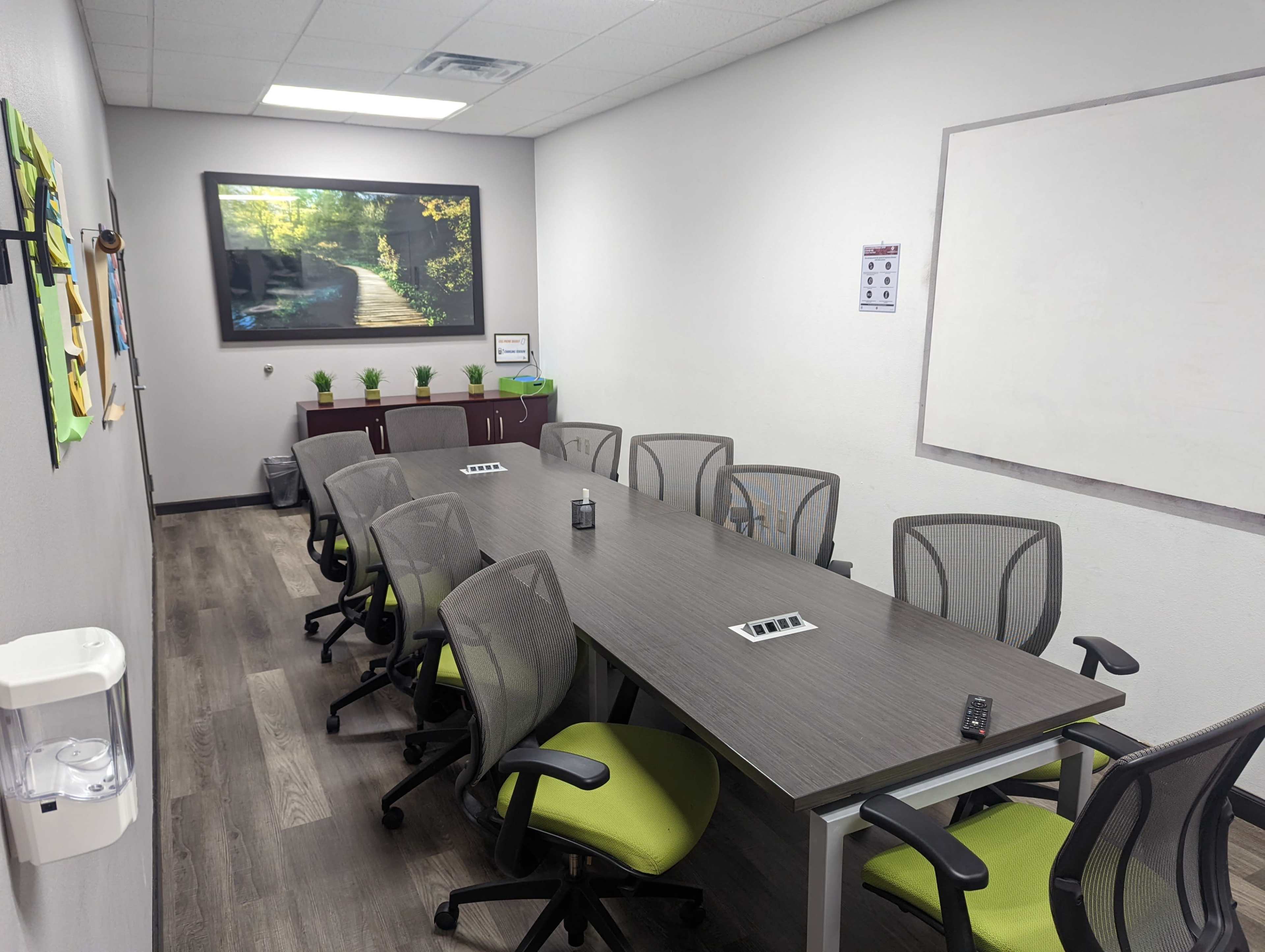 A conference room features a large table surrounded by ten chairs, with a wall-mounted screen displaying a nature scene and potted plants along the windowsill.