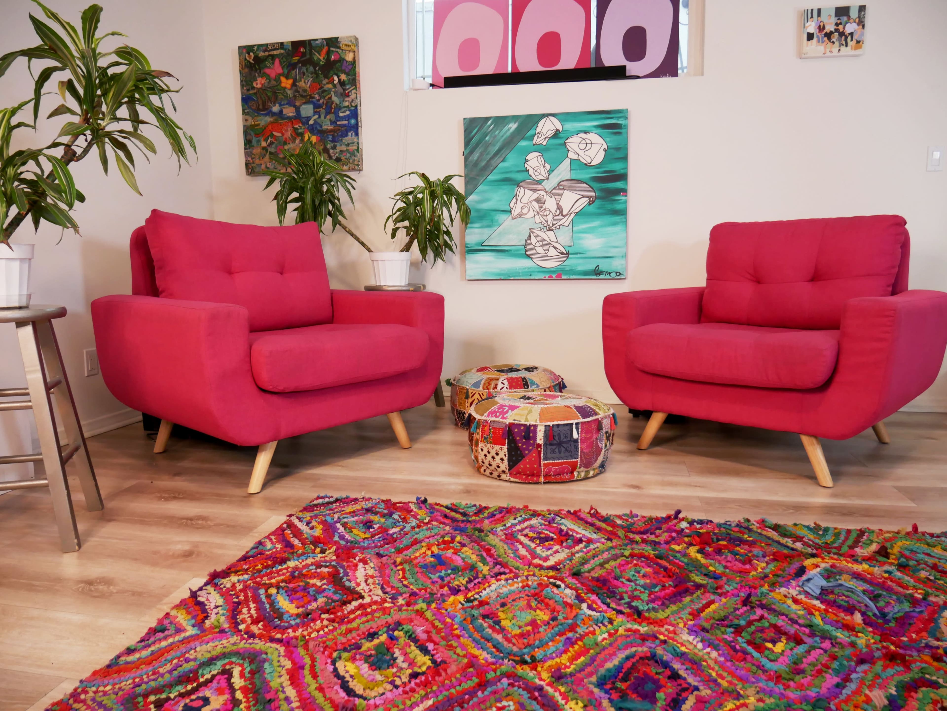 A brightly colored living room with two pink armchairs, a patterned pouf, a vibrant area rug, and various plants and artworks on the walls.