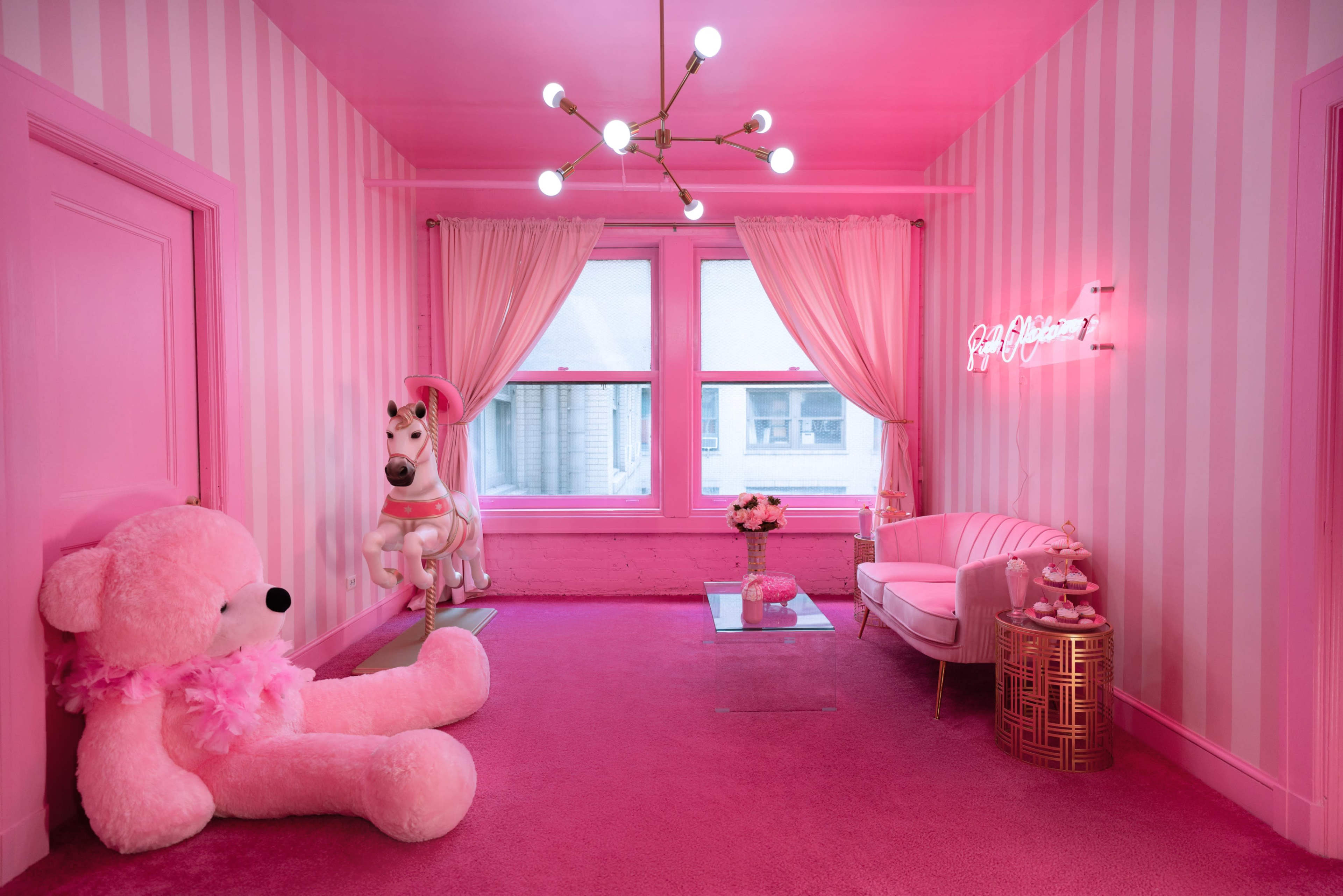 The room features pink striped walls, a large pink teddy bear, a unicorn statue, a pink sofa, and a neon sign that reads "Barbie Dreamhouse."