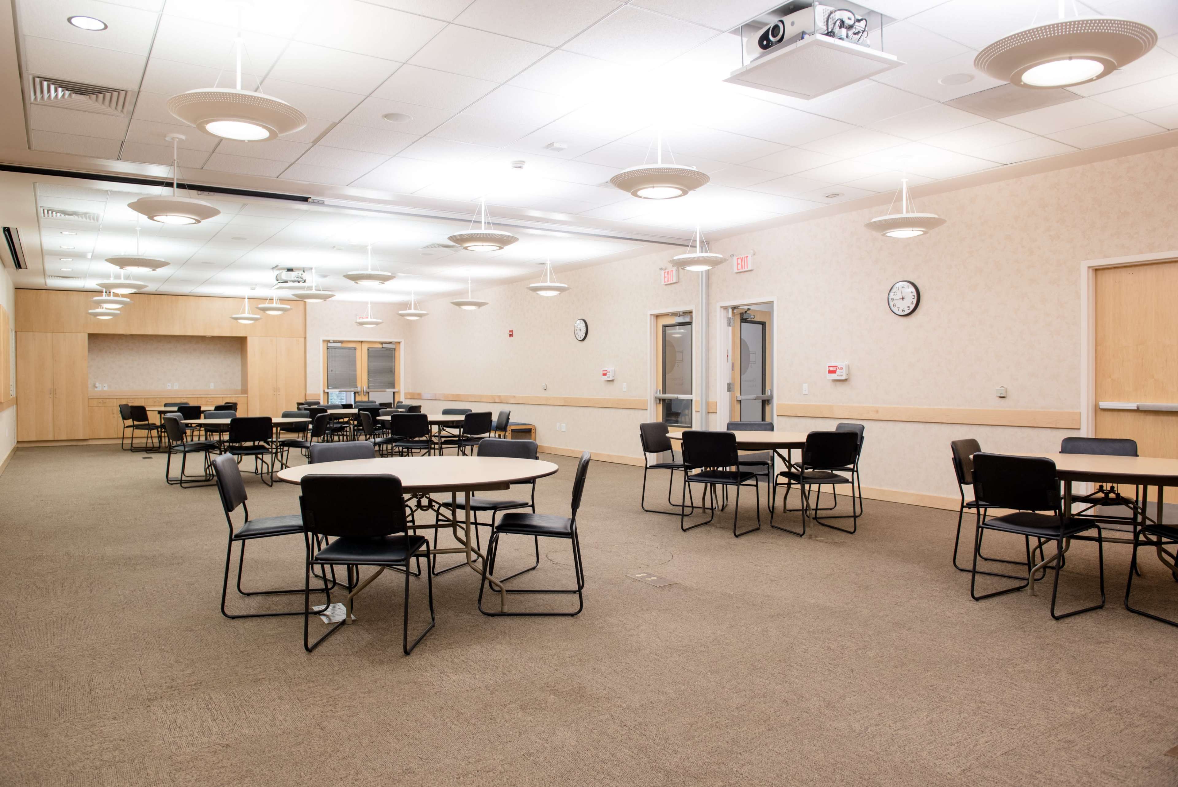 A spacious room with several round tables and black chairs, featuring overhead lighting and wall clocks.