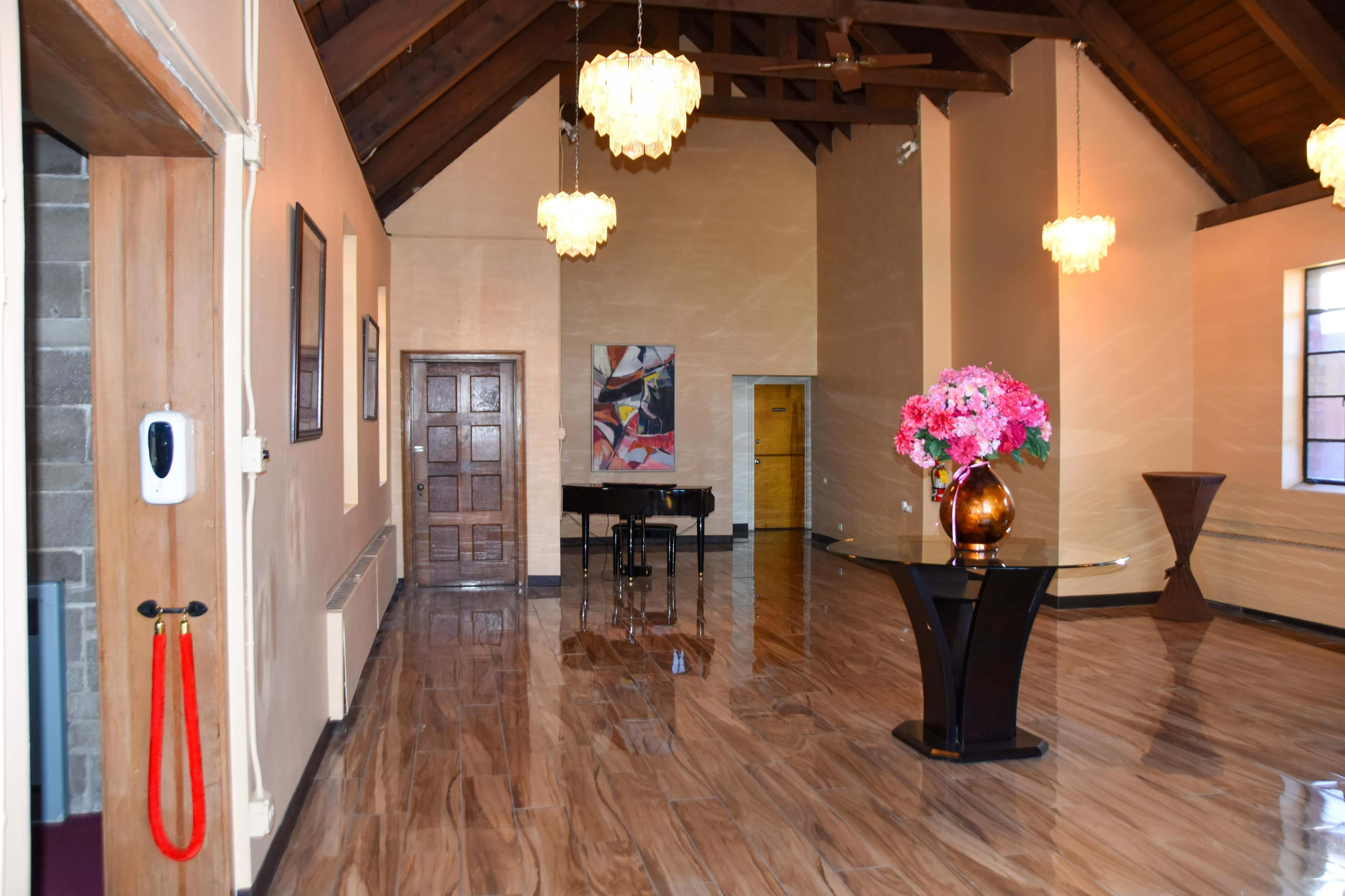 The image shows a spacious interior with high wooden ceilings, polished marble floors, a grand piano, and decorative lighting, along with a floral arrangement on a pedestal.