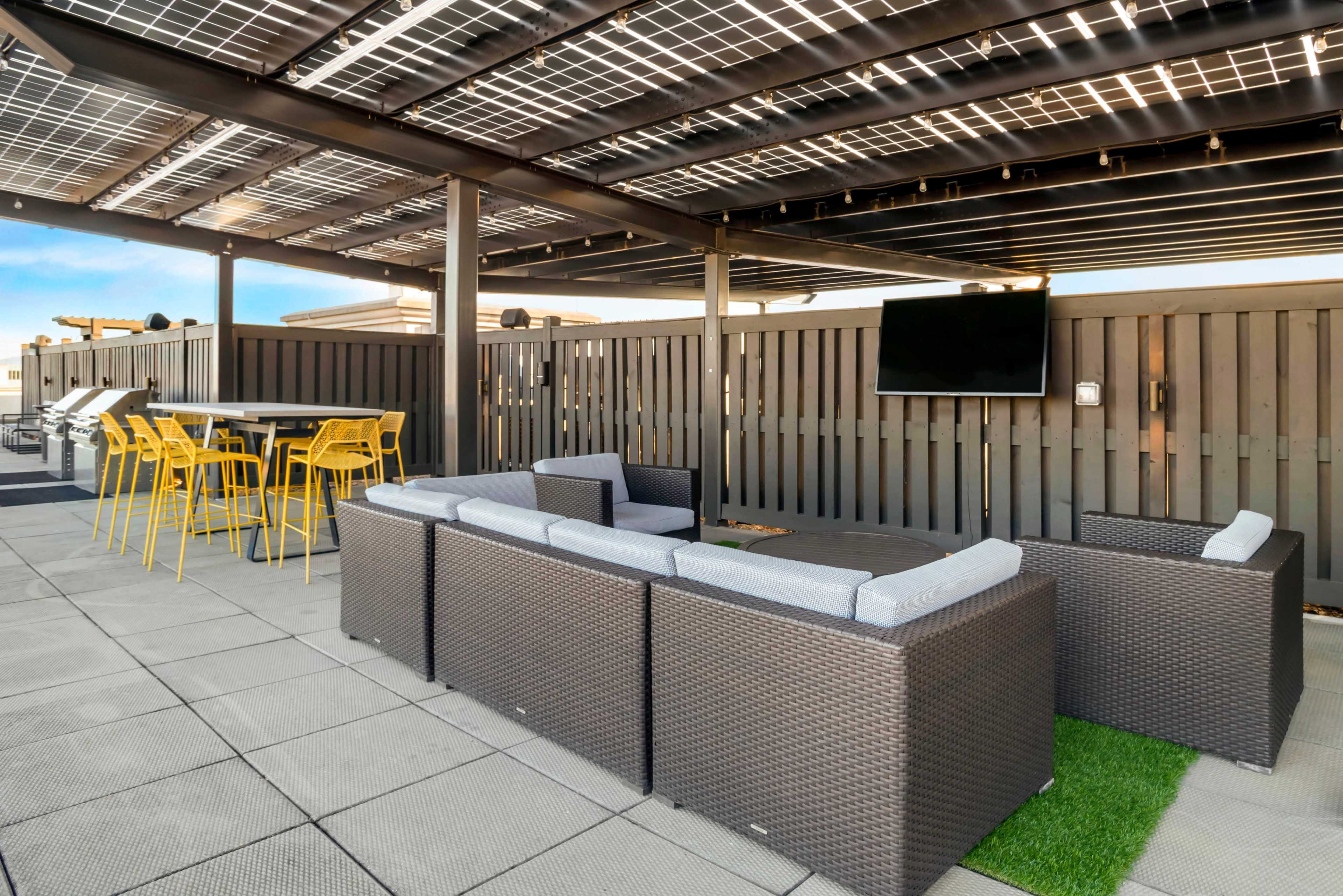 A rooftop seating area features a sectional couch, a table, bright yellow stools, and a mounted television under a pergola with solar panels.