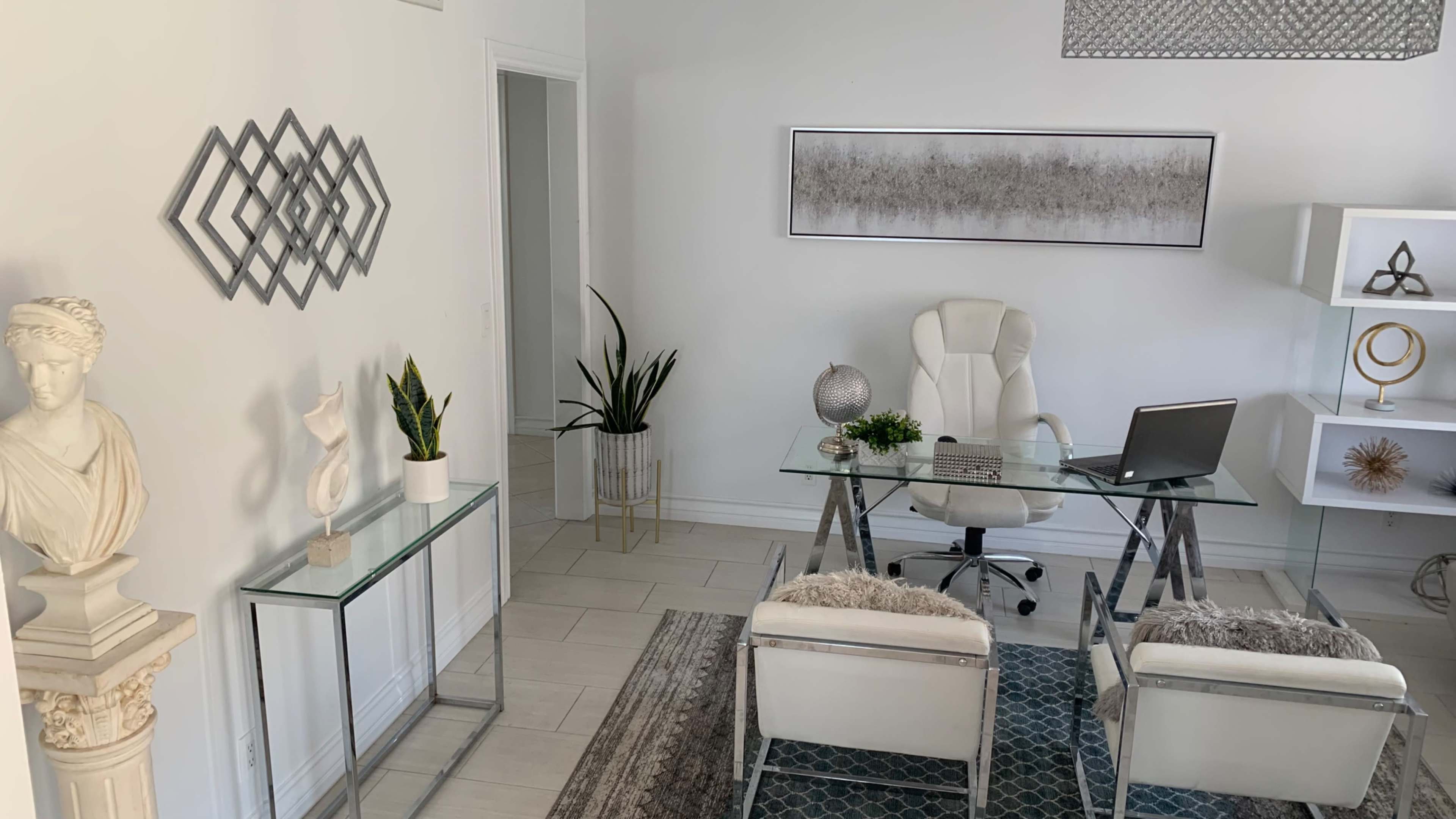 A modern office space features a glass desk, white chairs, decorative plants, and artwork on the walls.