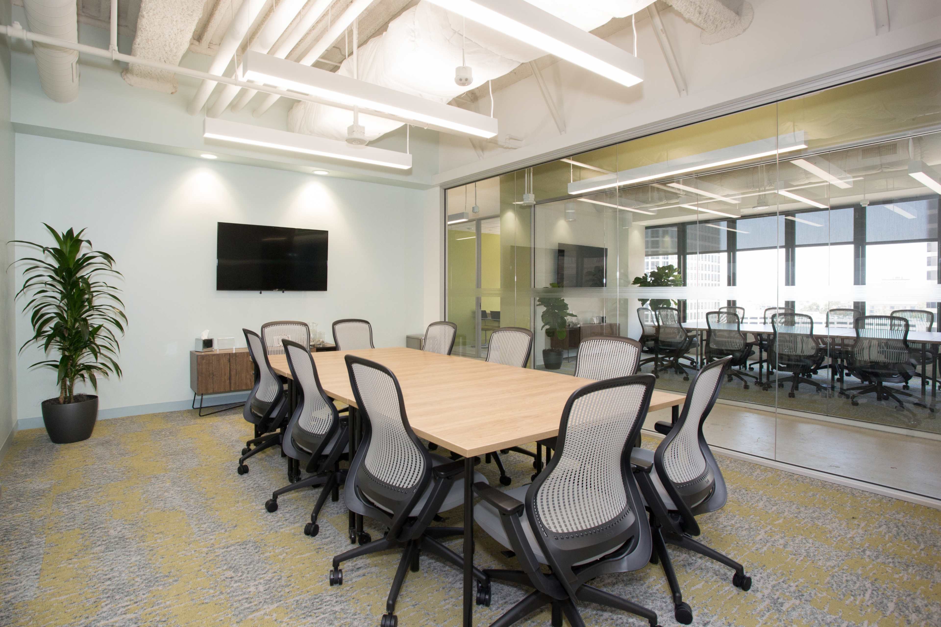 A modern conference room features a large wooden table surrounded by ergonomic chairs, with glass walls and a wall-mounted TV.