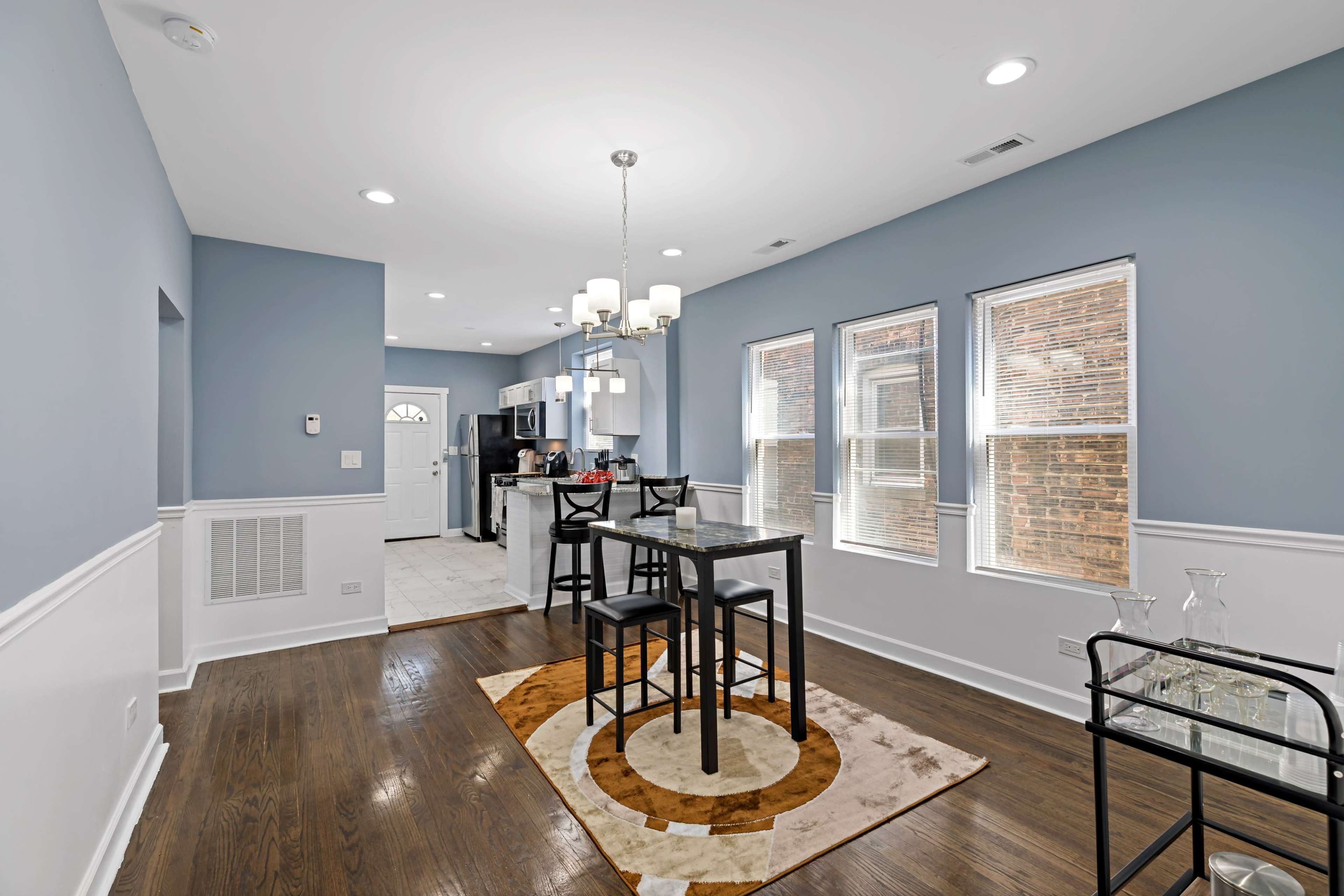 A kitchen and dining area with a table and four chairs on a patterned rug, with windows allowing natural light to filter in.