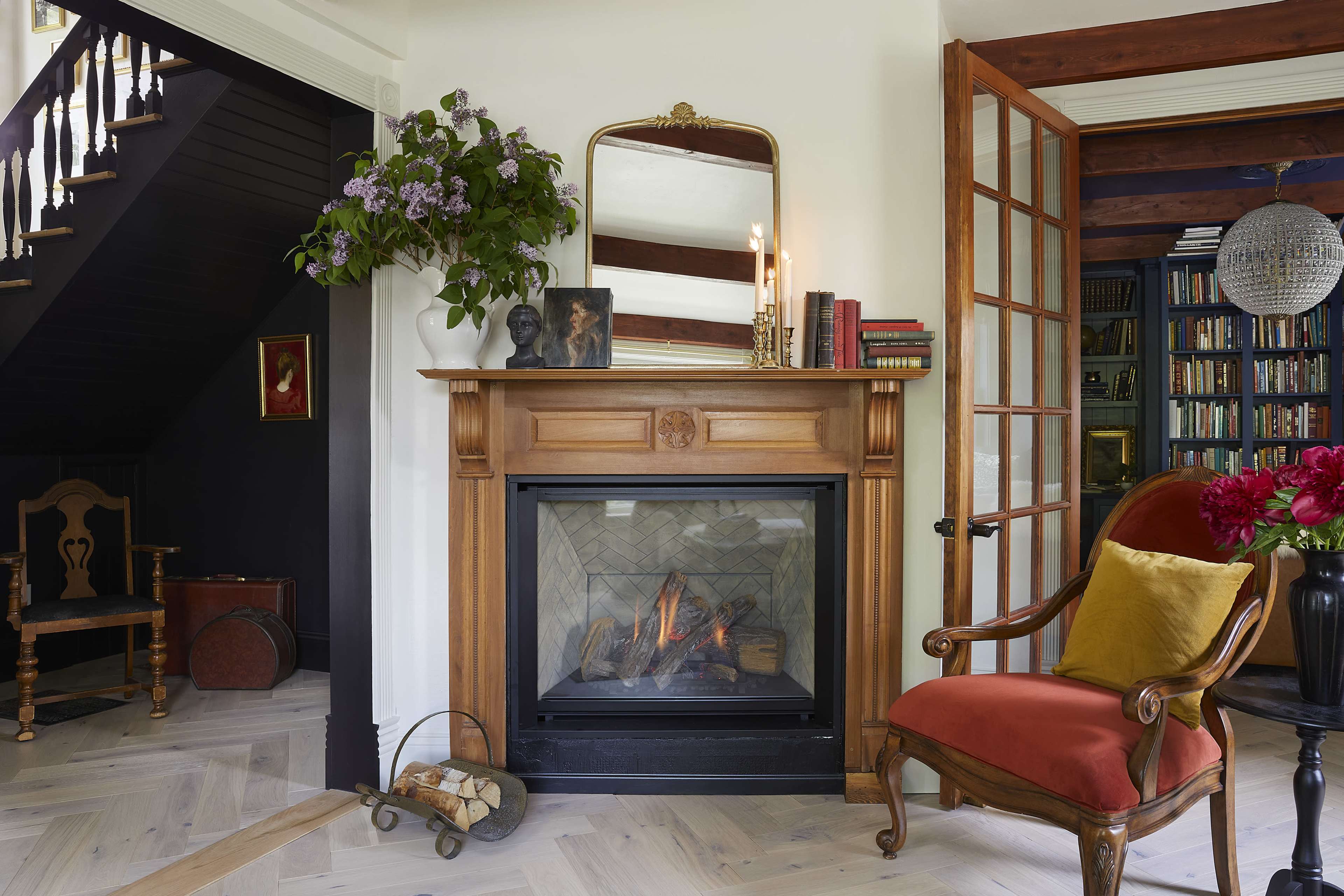 The image shows a cozy living room with a wooden fireplace, a large mirror above it, an armchair with red upholstery, a staircase, and bookshelves in the background.