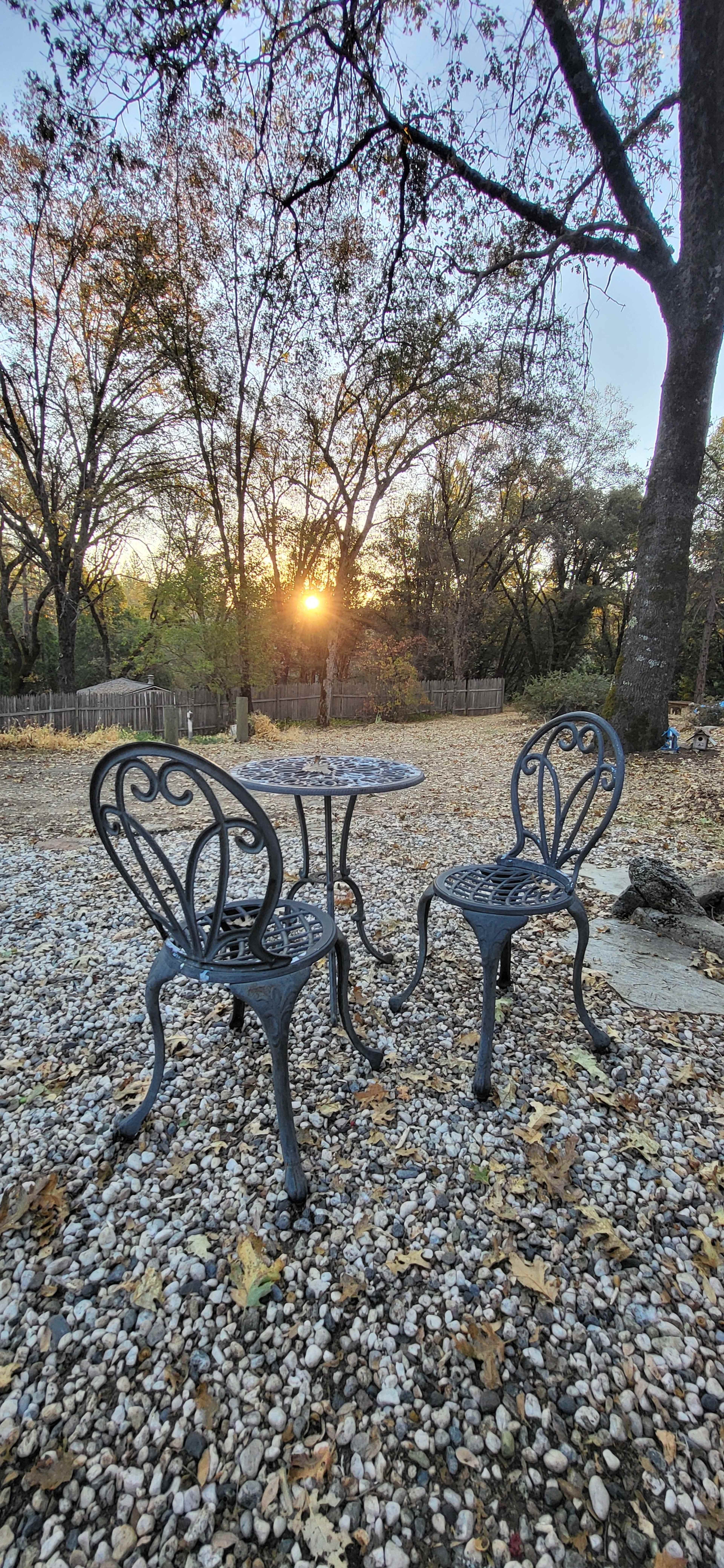 A small metal table and two chairs sit on a gravel surface with trees in the background and the sun setting on the horizon.