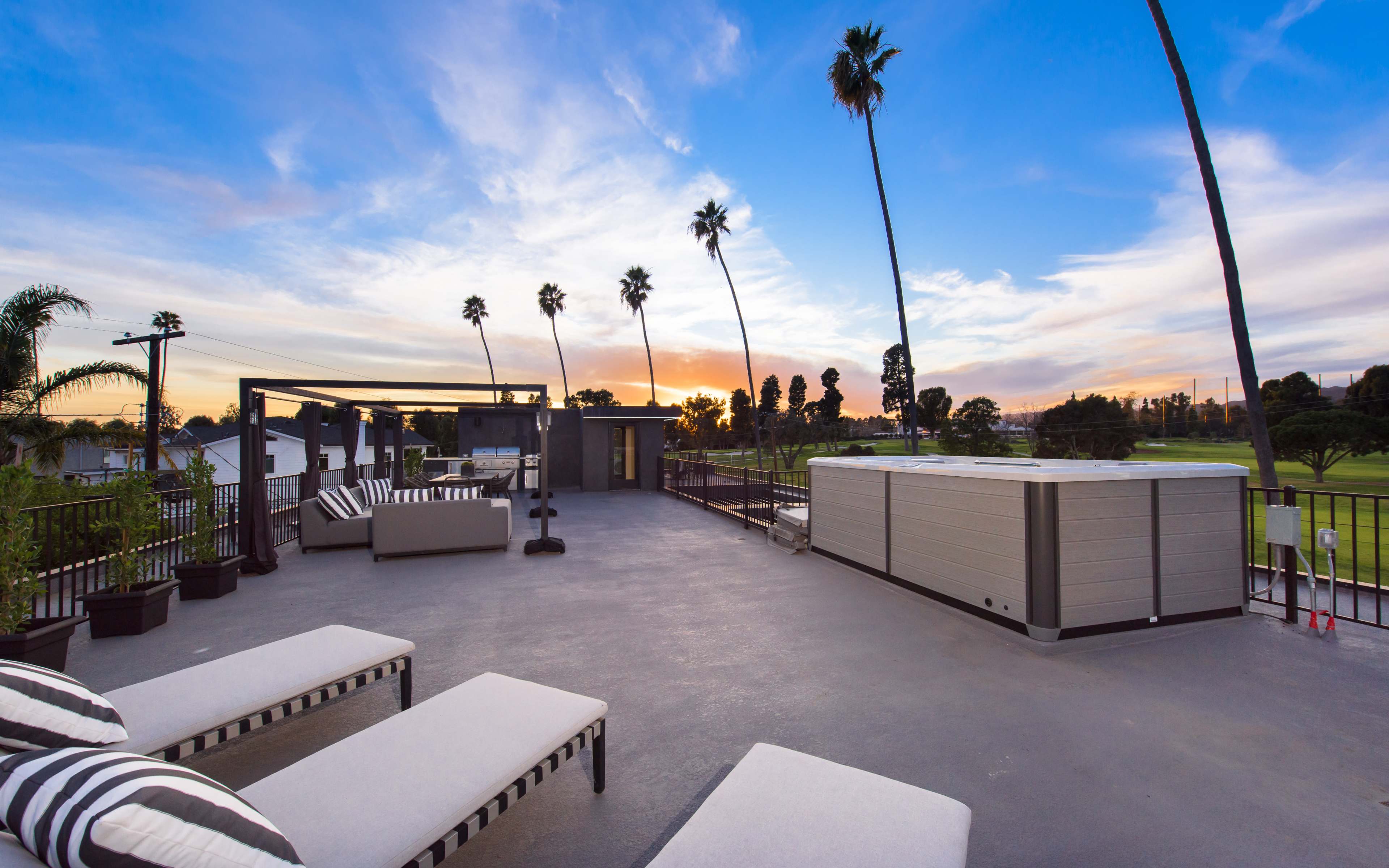 A rooftop patio features lounge chairs and a hot tub, surrounded by palm trees under a colorful sunset.