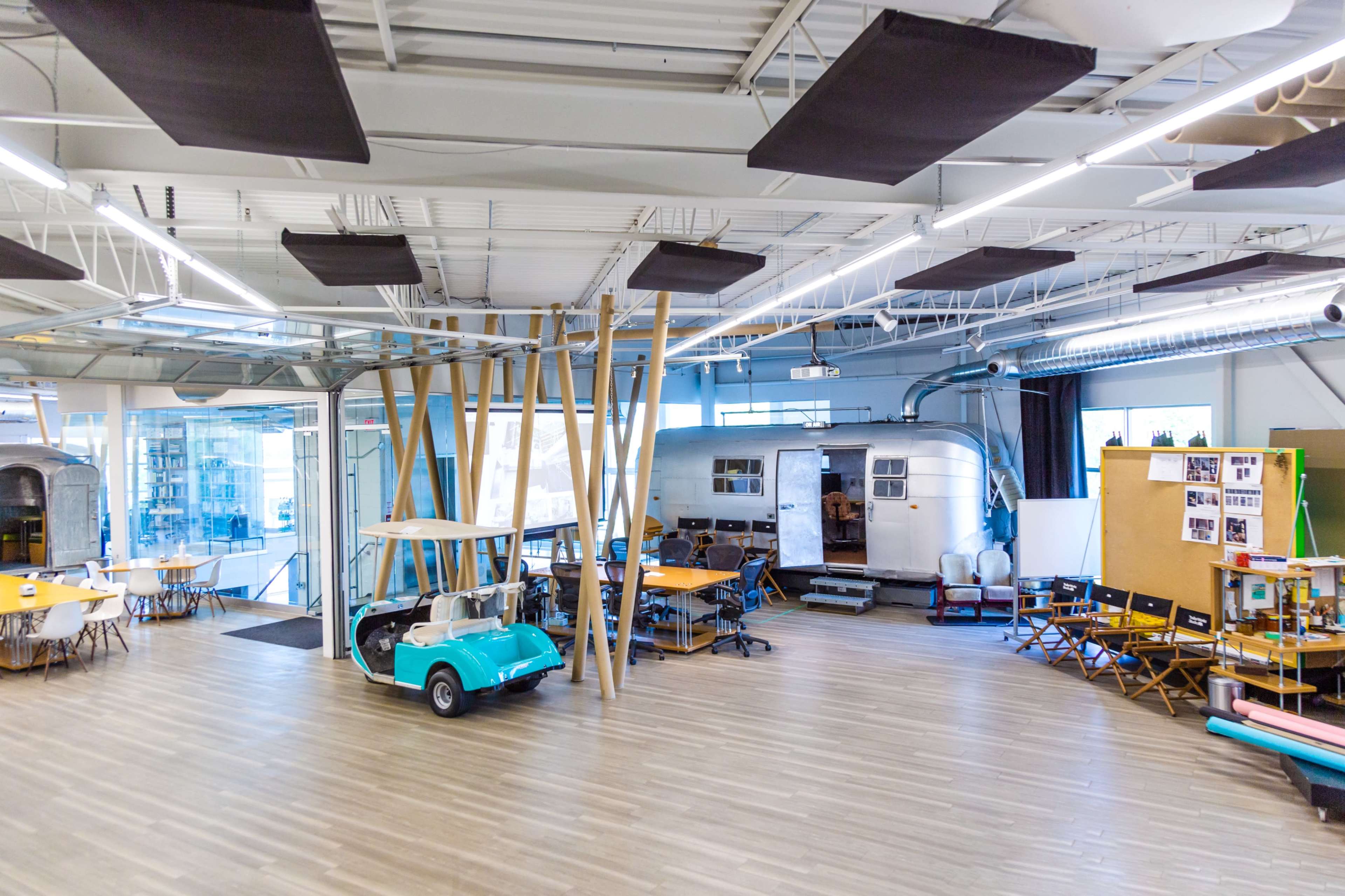 The image shows an open office space featuring a retro camper, tables, chairs, and a golf cart, with large windows allowing natural light to enter.