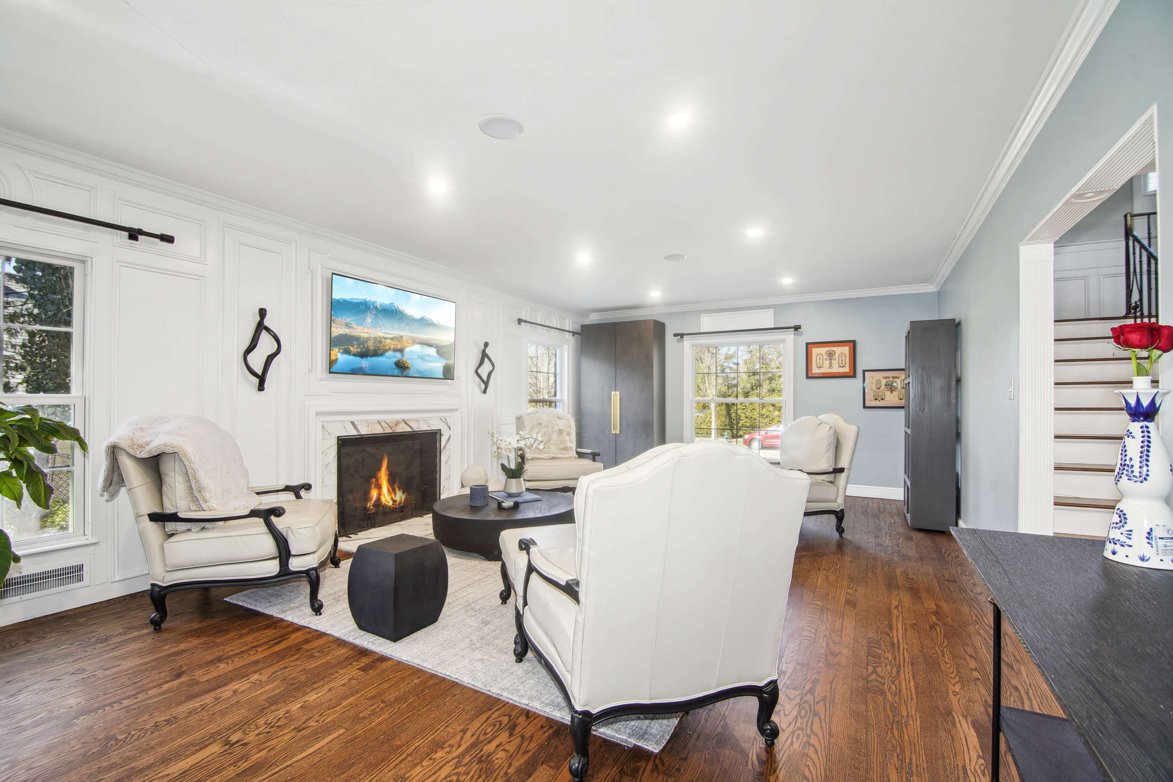 A modern living room features two white armchairs, a black coffee table, a fireplace, and a mounted television, with large windows allowing natural light in.