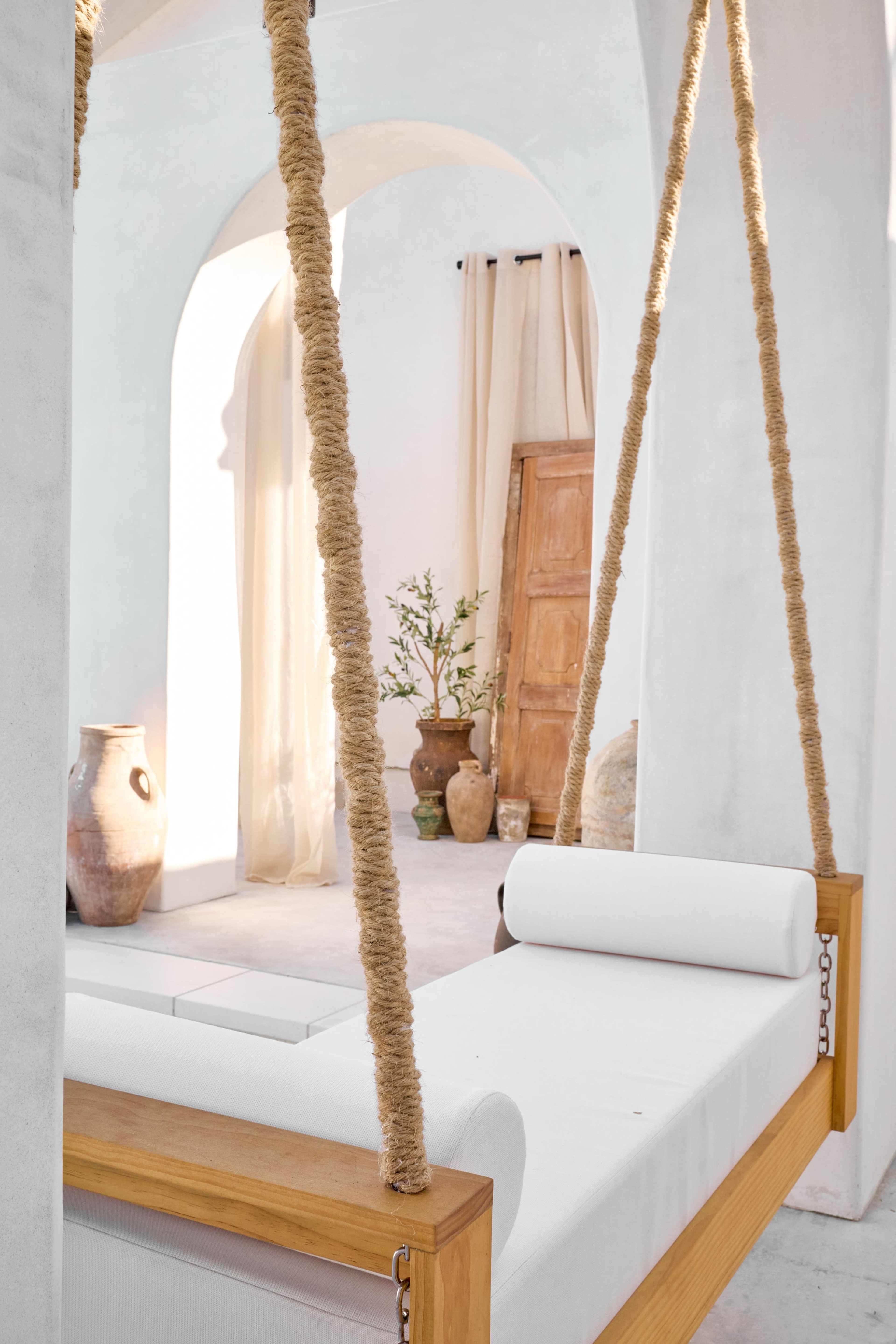 A swing bed made of wood and white fabric is suspended by thick ropes in a minimalist interior featuring white walls and earthy decor.