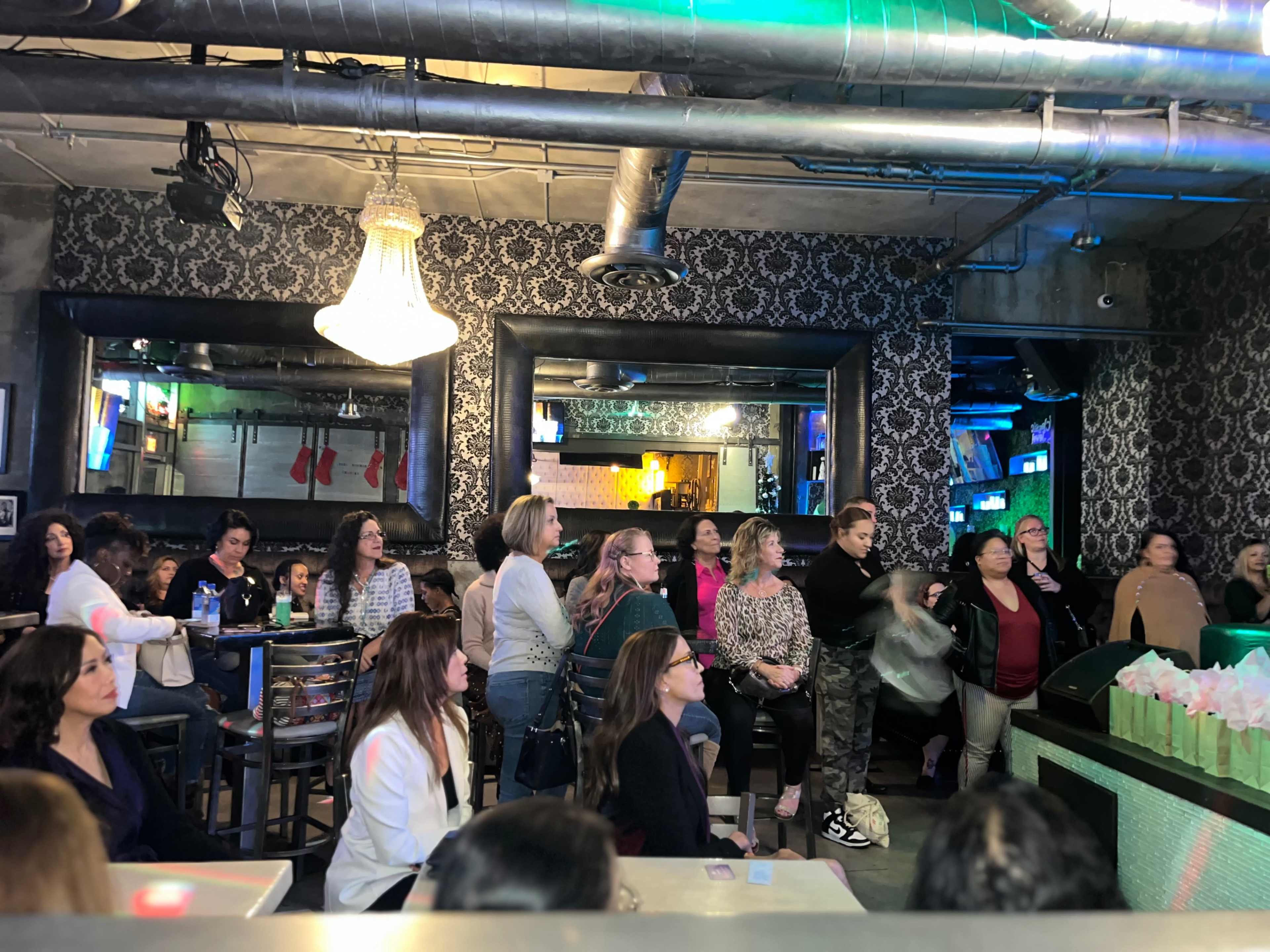 A diverse group of women is gathered in a dimly lit bar, watching a performance with several mirrors and decorations in the background.