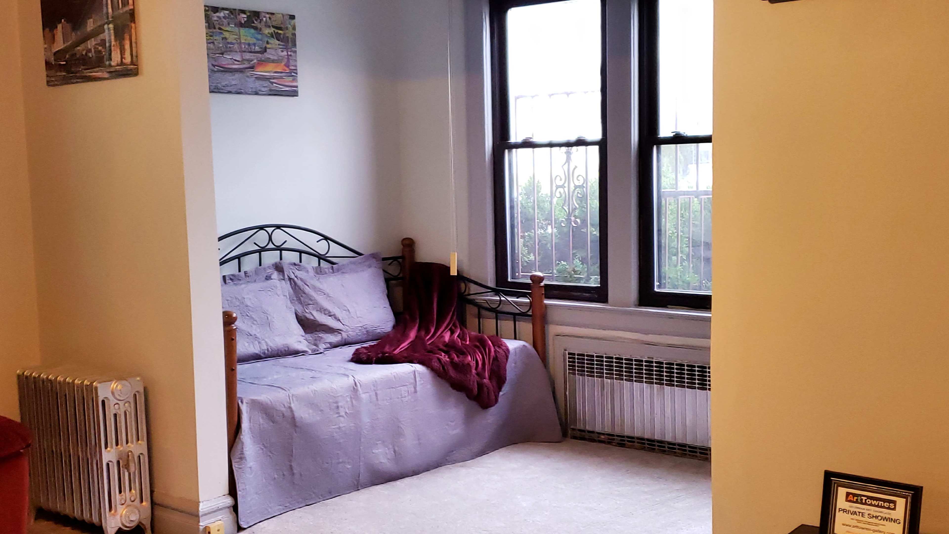 The image shows a corner of a room featuring a bed with gray bedding, a maroon blanket draped over it, a large window with metal bars, and a radiator nearby.