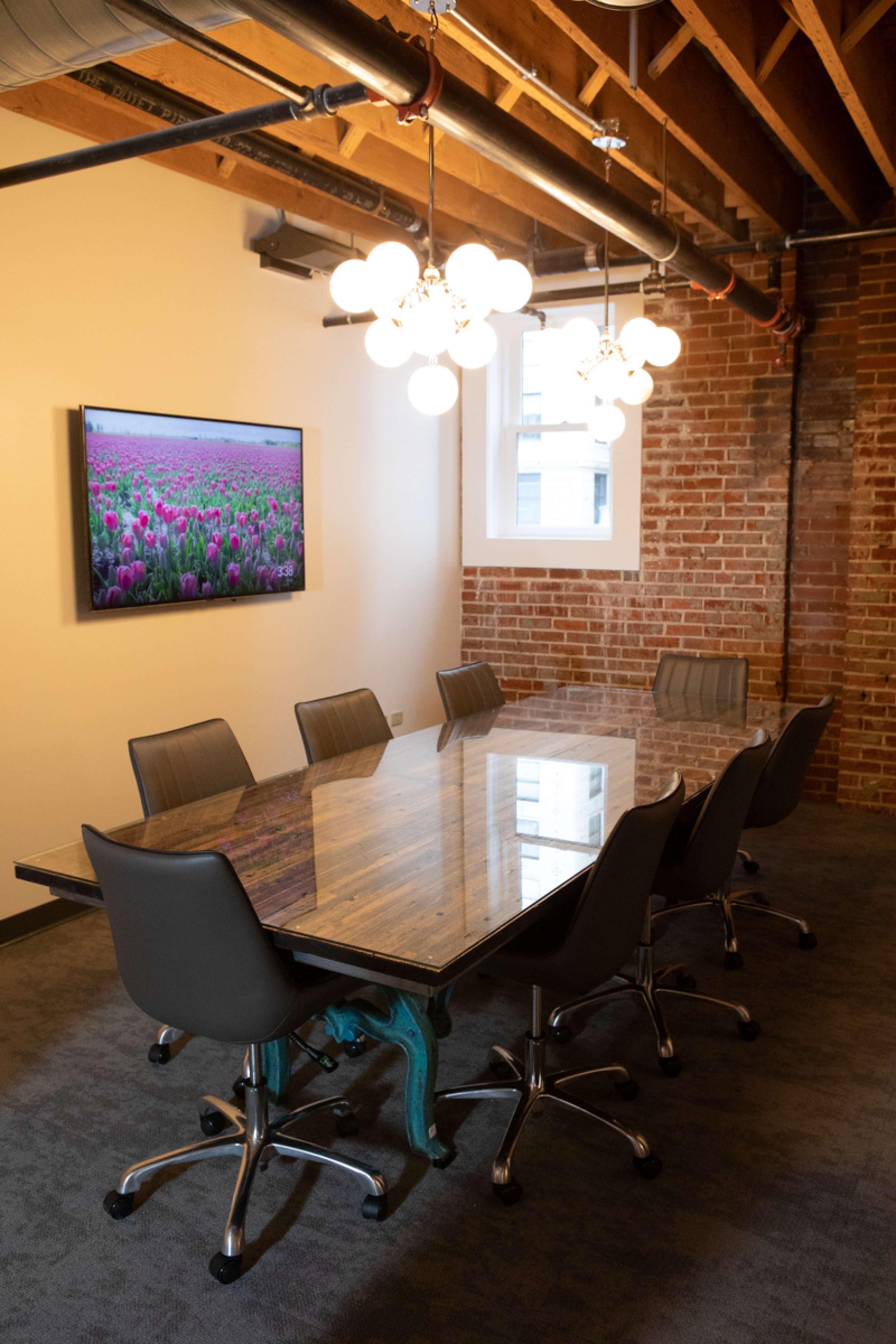 A conference room features a large glass table surrounded by six rolling ergonomic chairs and a television mounted on the wall displaying a floral image.